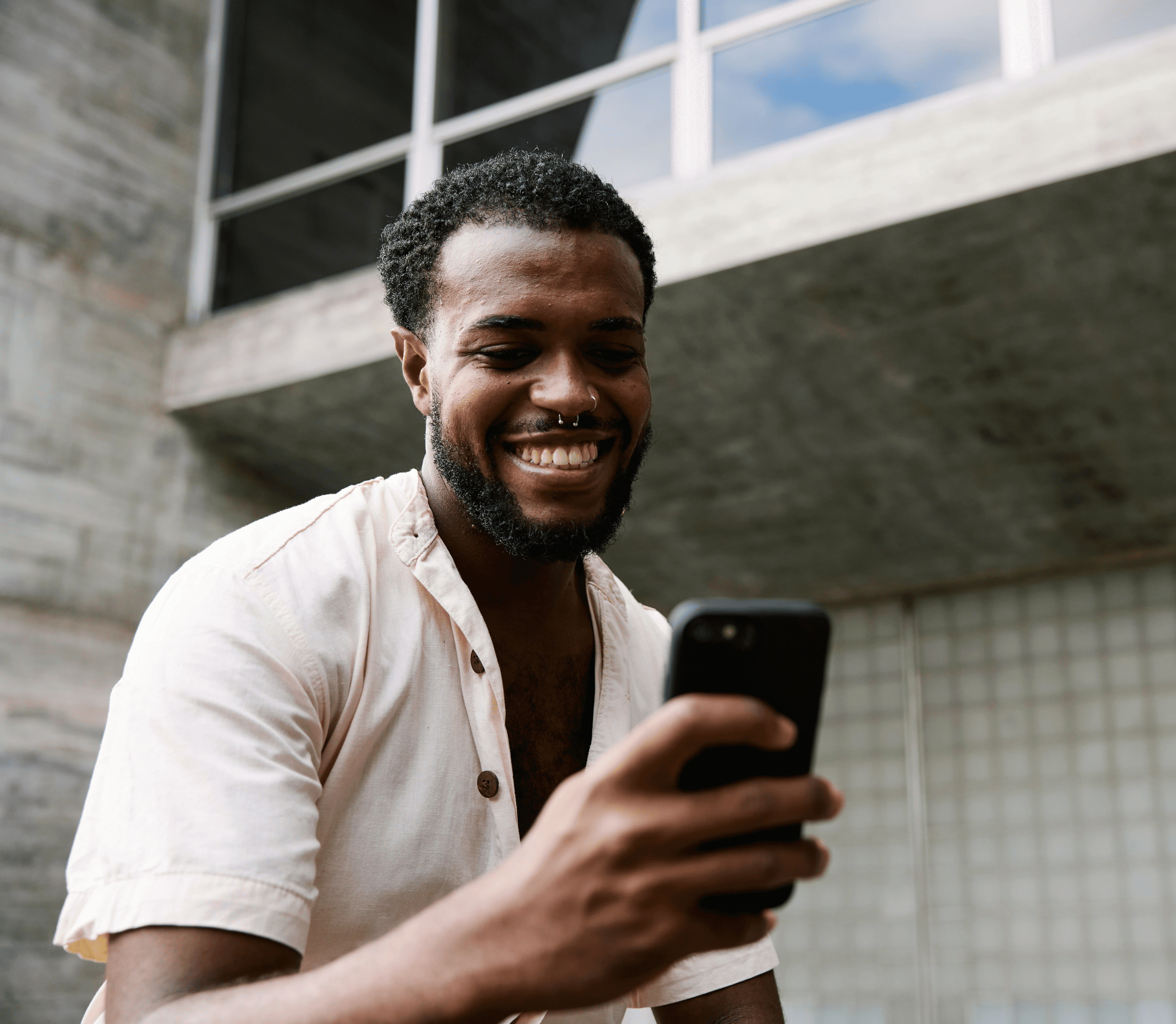 A smiling man holds a smartphone, enjoying a moment outdoors near a modern building.
