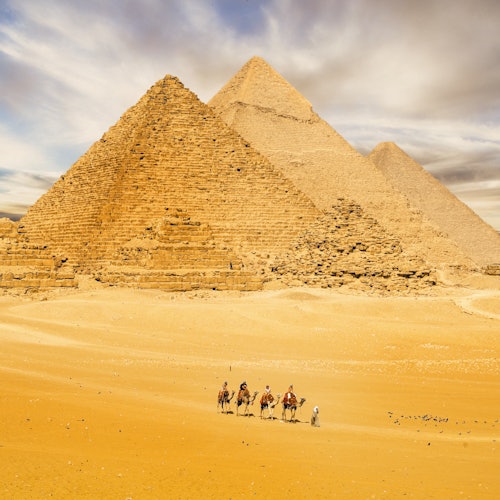 Three pyramids in a desert with four people riding camels on the sandy terrain under a partly cloudy sky.