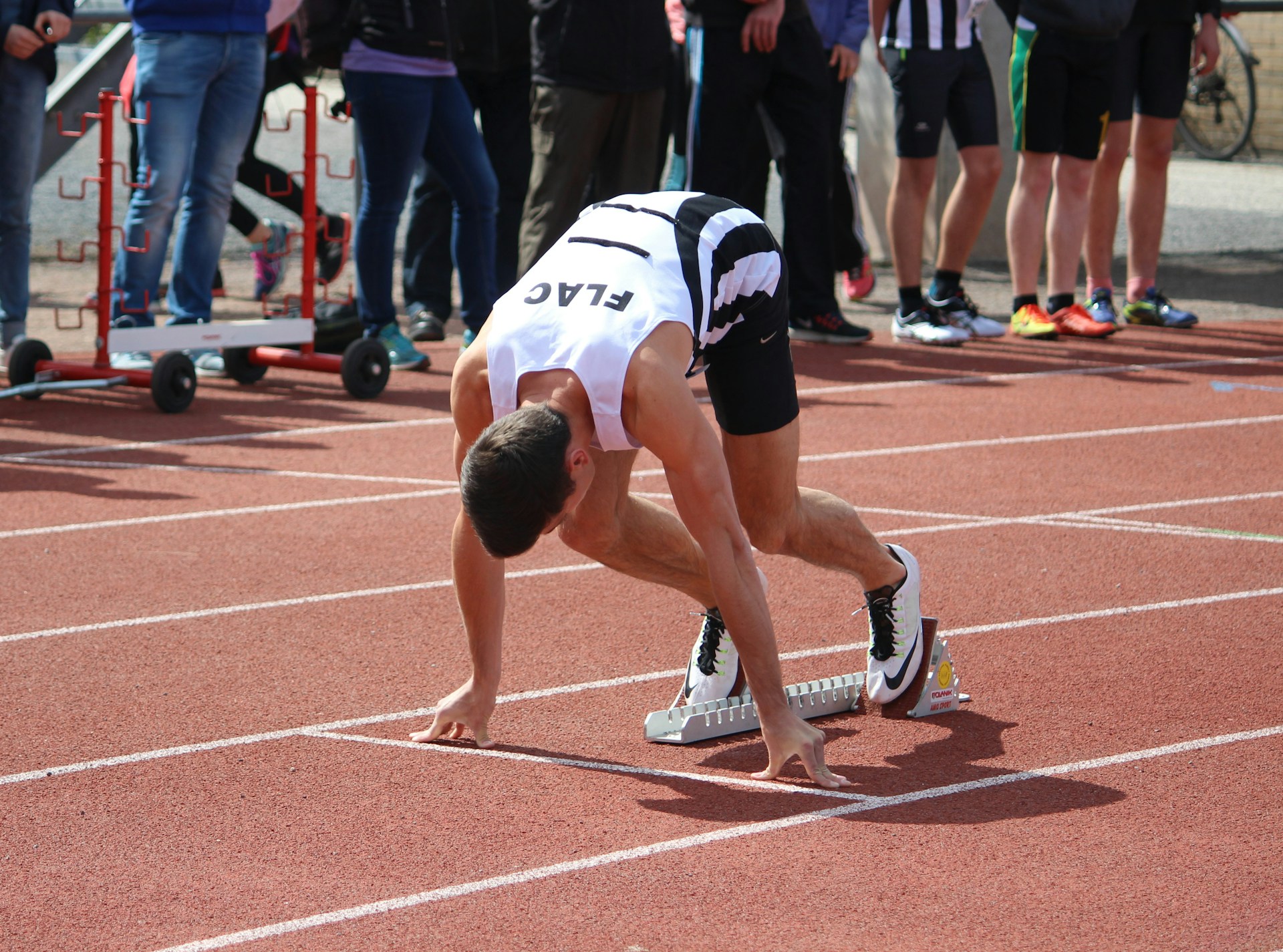 A male track athlete crouches in the starting blocks on a red track, head down in preparation for a sprint.