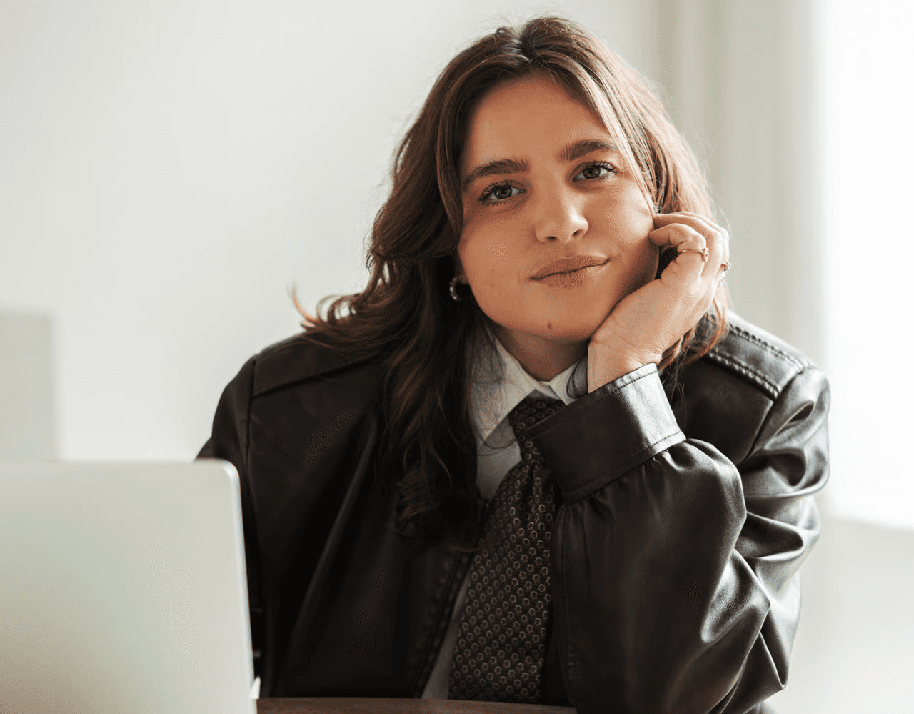 A person with short brown hair looks confidently at the camera, wearing a light pink blazer and white shirt. Soft lighting and plant in the background add a calming tone.