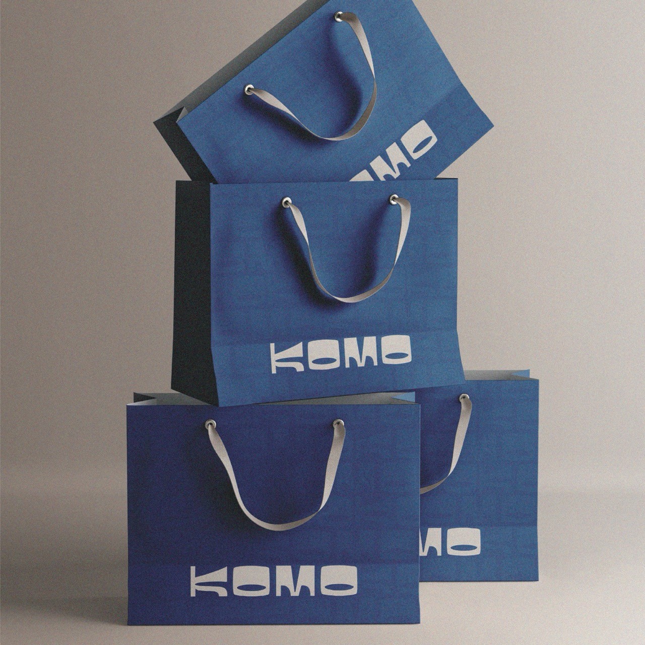 Komo- Four stacked blue shopping bags with white handles and the bold text "KOMO" printed on them. The arrangement forms an artistic, balanced display.