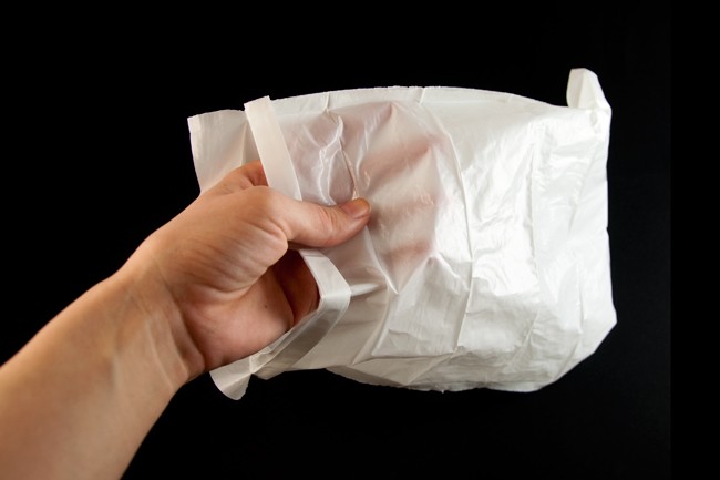 A hand is holding a white, crumpled plastic bag against a black background. The hand grips the bag near the top, pulling it slightly open. The bag’s surface is wrinkled and creased, indicating it has been handled. The hand’s skin tone is light, and the background is uniformly dark, providing a stark contrast to the white bag.