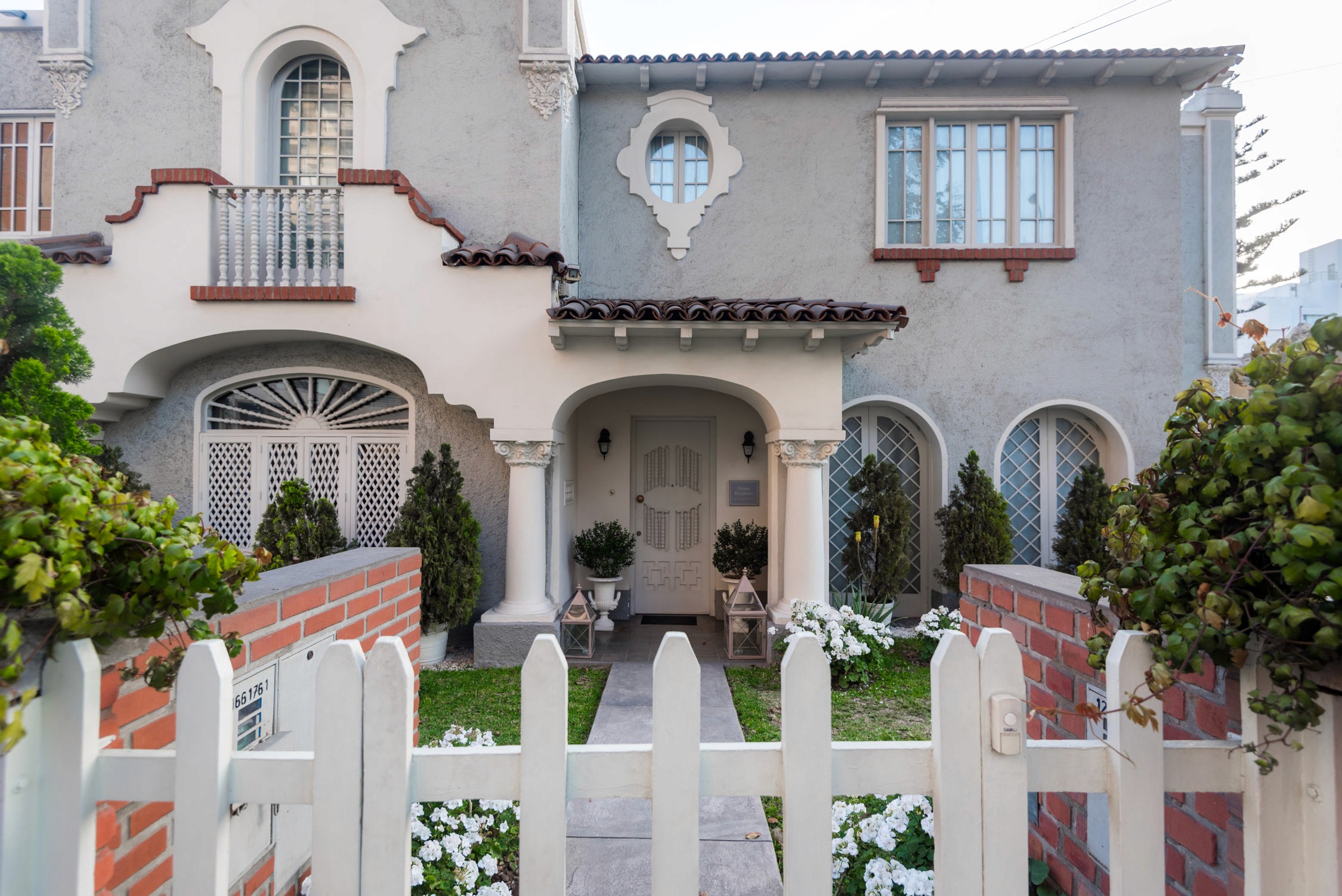 A gray stone villa with arched windows and a white picket fence decorated with white flowers in front of it.