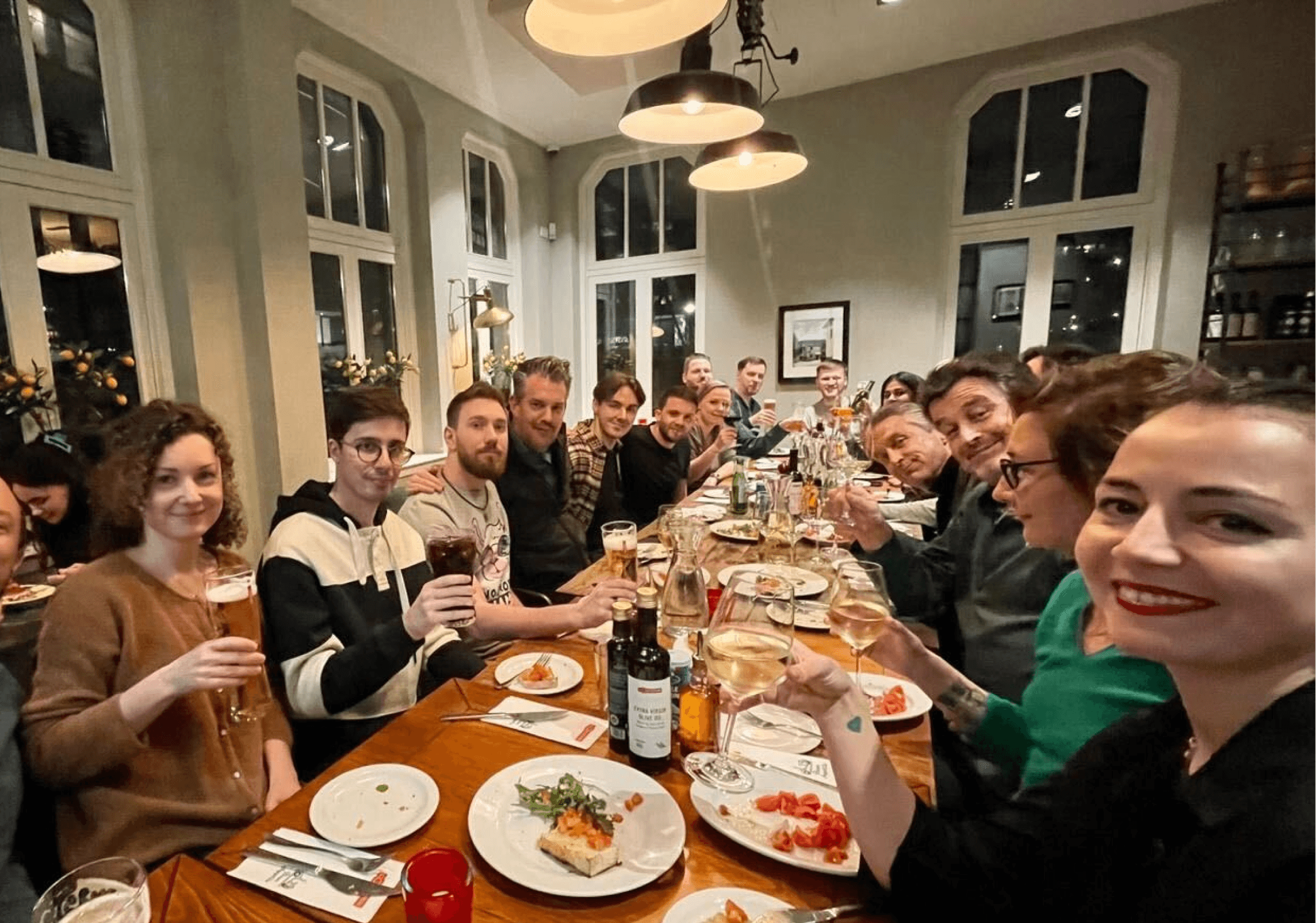 Un grupo grande de compañeros de trabajo sonríe alrededor de una larga mesa de restaurante, compartiendo la cena y brindando con sus copas bajo unas cálidas luces colgantes.