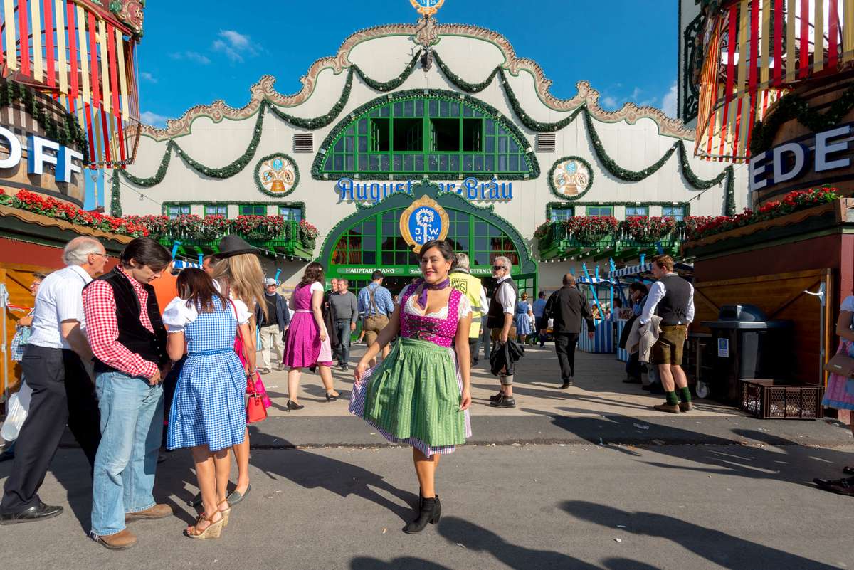 People in colorful costumes and traditional attire walk past a large "Hüpfburg" bouncy castle at a fairground.