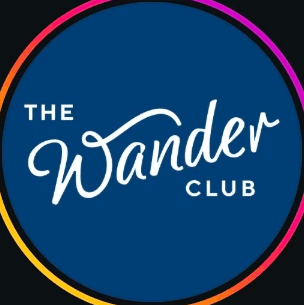 A woman with blonde bob hair in a grey coat and beige turtleneck stands against a neutral backdrop. She has a serious expression, conveying elegance.