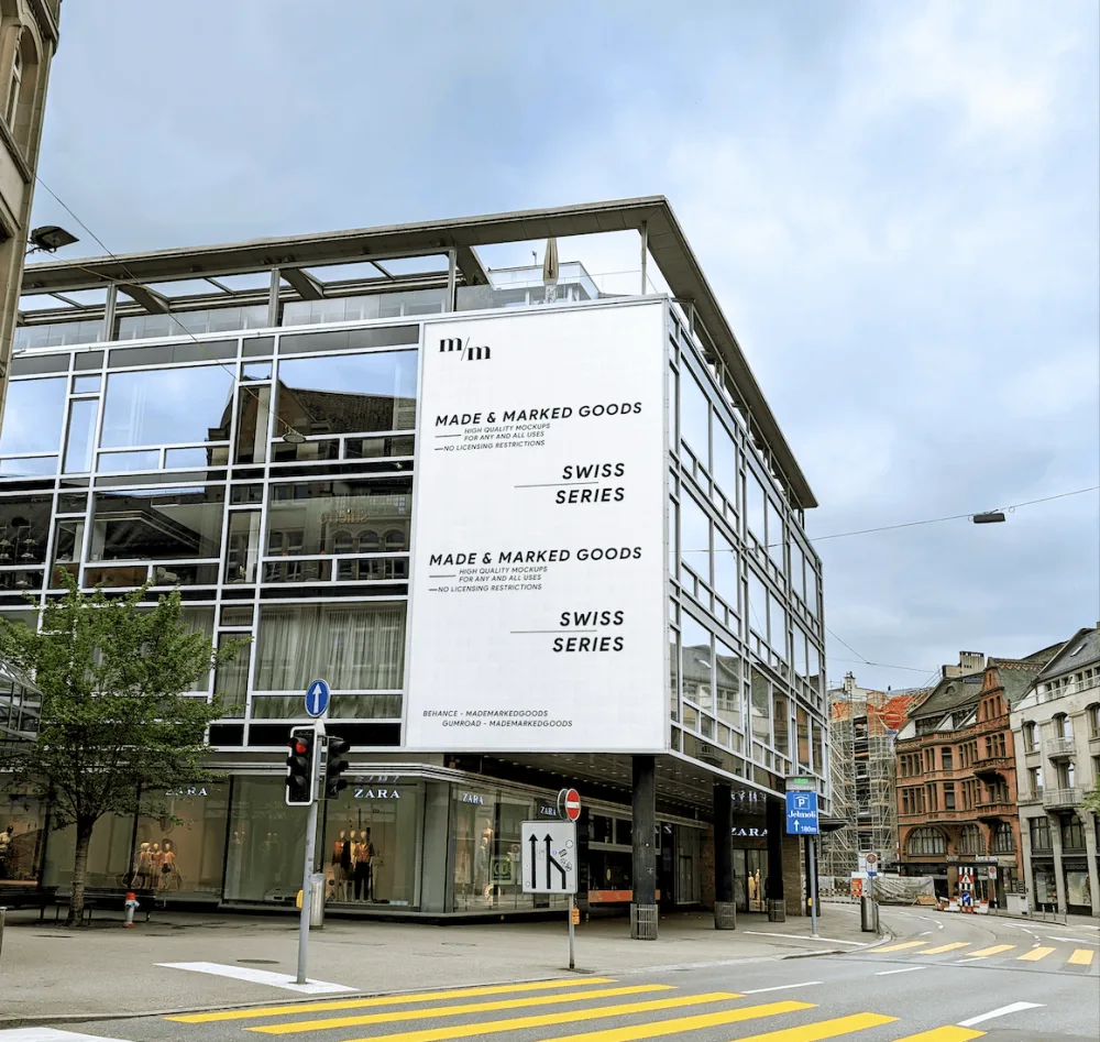 A modern glass building featuring a large white banner mockup with minimalist text, situated on a street corner with a Zara storefront below. The urban setting includes crosswalks, trees, and historic architecture in the background under an overcast sky.