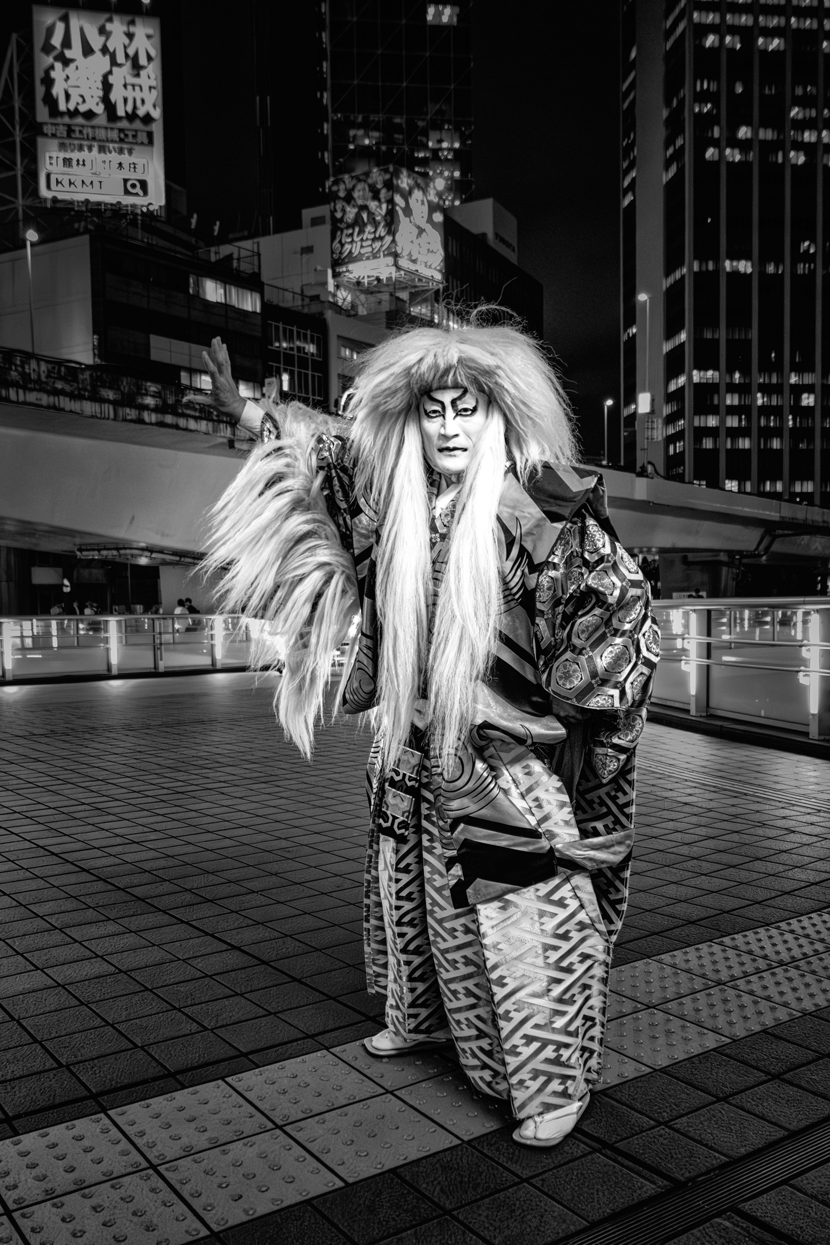 Black and white night portrait of a performer wearing elaborate costume and makeup standing in a city square.