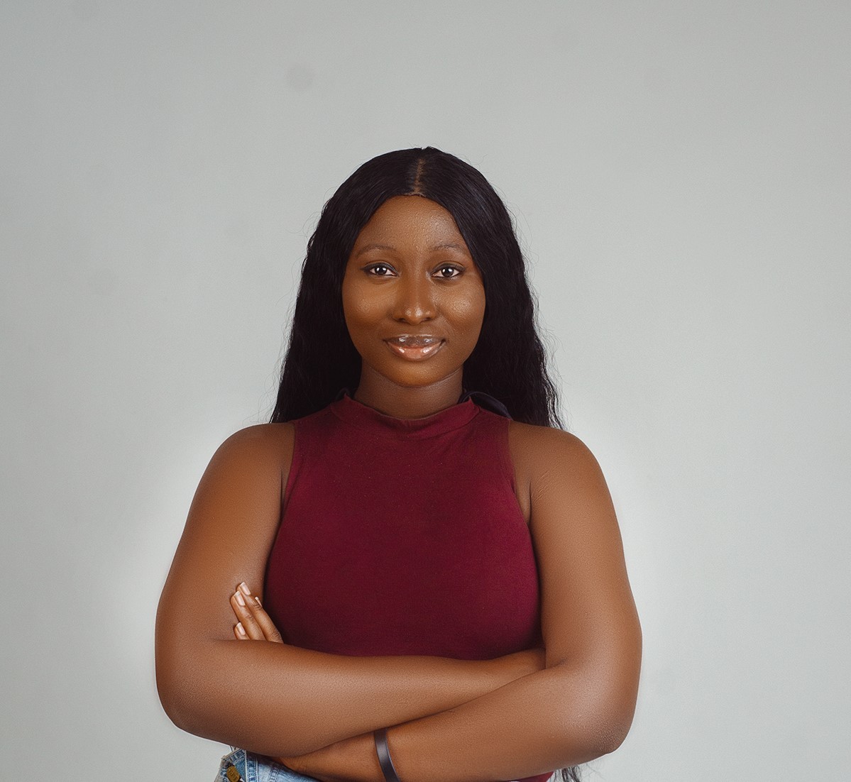 A confident woman with long black hair wearing a sleeveless burgundy top and blue jeans stands against a plain gray background, embodying the professional and approachable qualities ideal for a portfolio or profile image of a web or product designer.