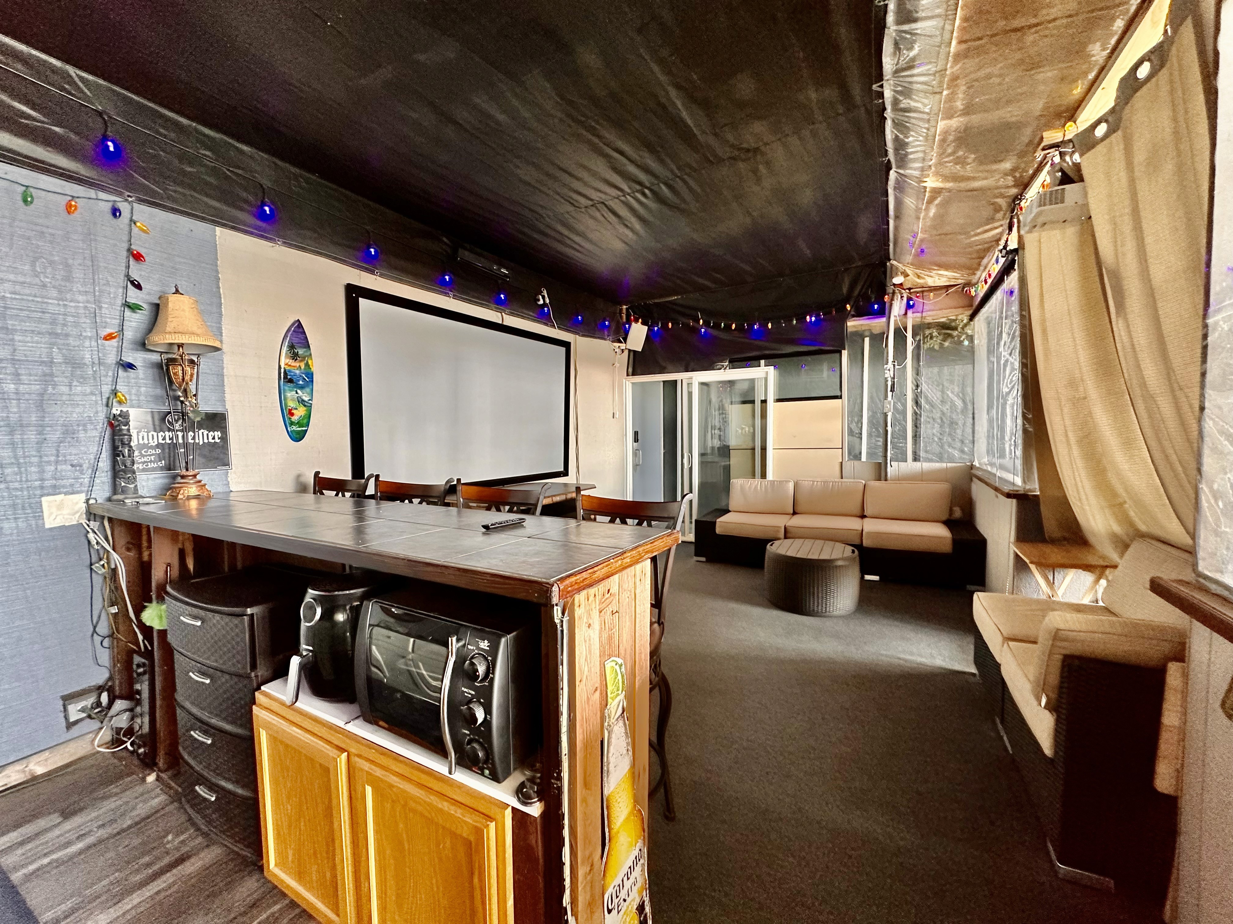A cozy basement entertainment room features a large projector screen, surrounded by comfortable beige sofas and upholstered seating, with string lights adding a warm ambiance and a well-equipped wooden bar counter topped with a lamp and a sound system.