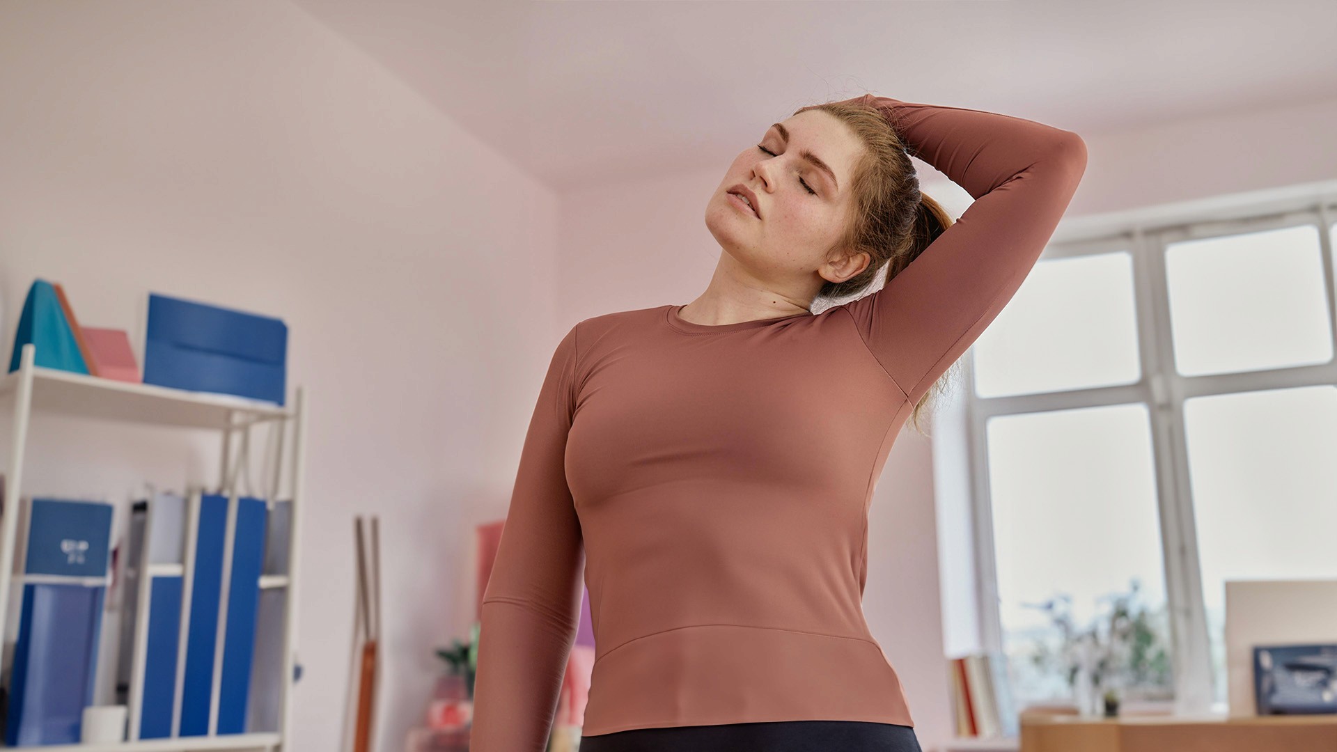 Junge Frau in einem Sportshirt führt eine seitliche Nacken-Dehnübung in einem hellen Raum aus, um ihre Muskulatur zu entspannen und ihre Flexibilität zu verbessern.