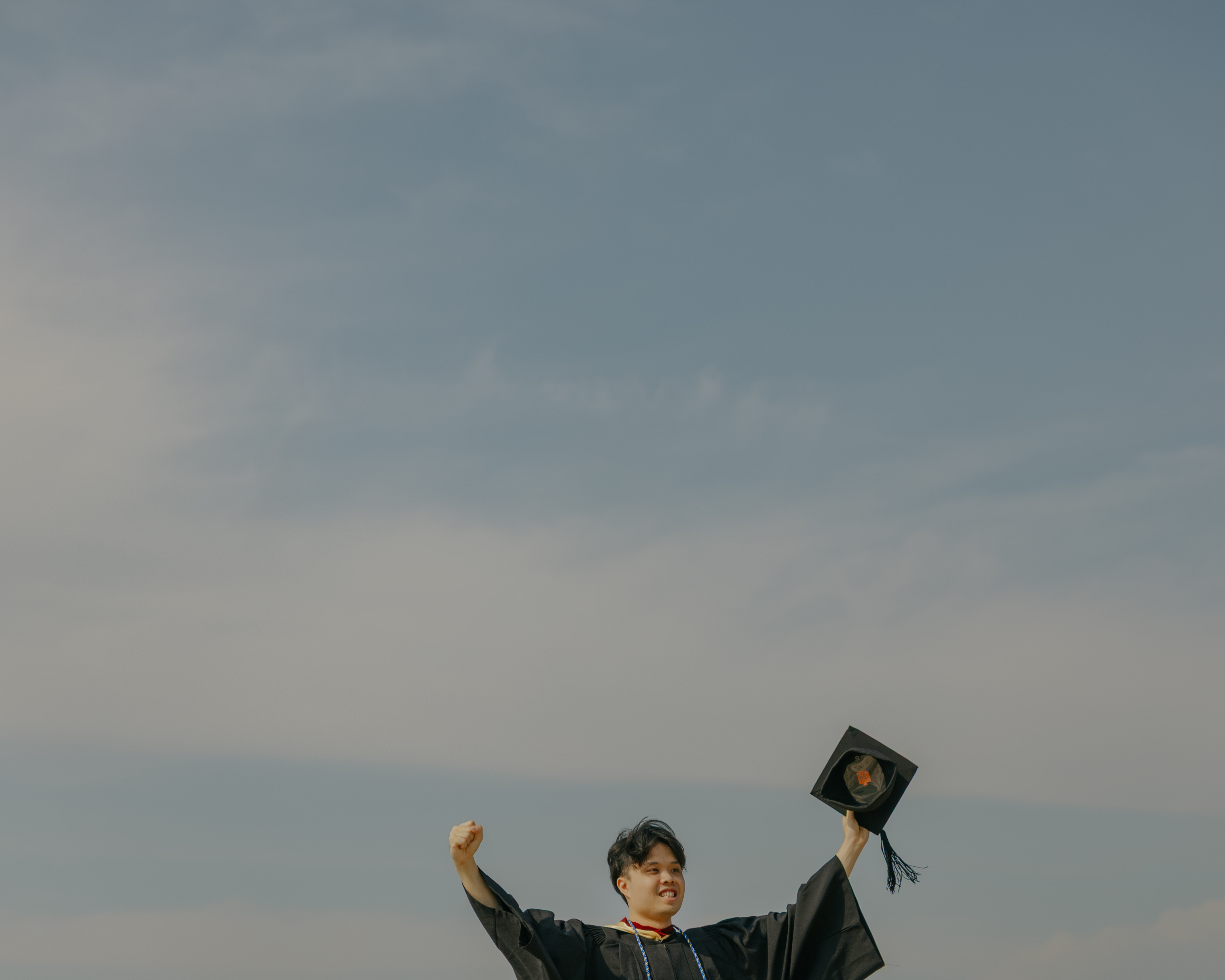Person celebrating outdoors, arms raised joyfully, holding a hat against a clear blue sky.