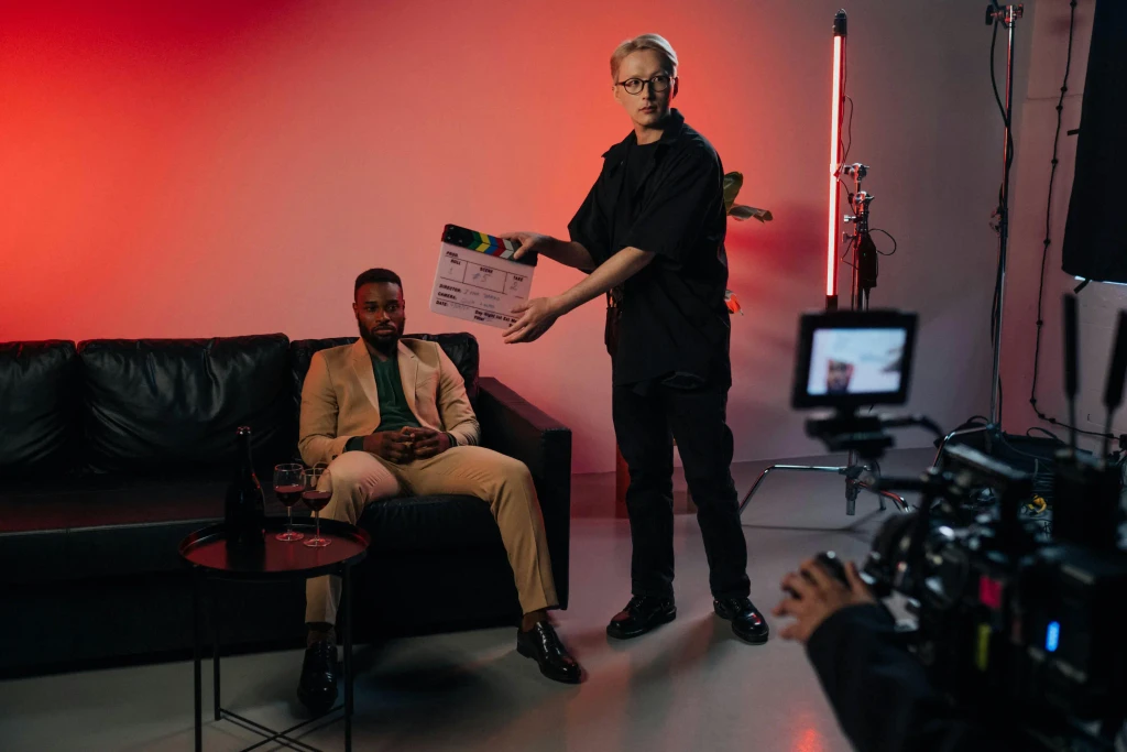 In a red-lit studio, a crew member holds a clapperboard before a man in a tan suit on a couch, seen behind a camera.