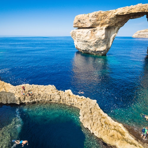 People swimming and sunbathing on rocky formations by a natural arch over clear blue sea under a clear sky.