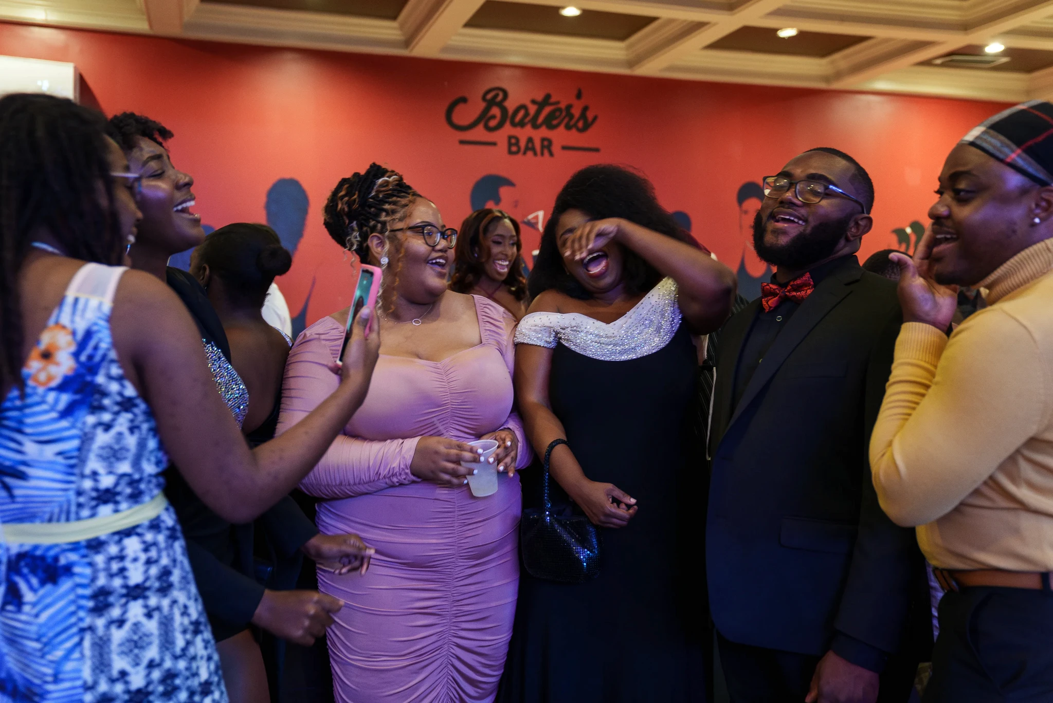 A group of people dressed in formal attire is laughing and enjoying themselves at an event. They are standing in front of a red wall with Bates Bar written on it.