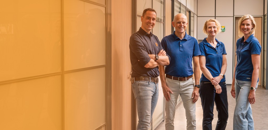 A group of four people, two men and two women, stand together smiling in a bright, modern office hallway, dressed in casual professional attire.
