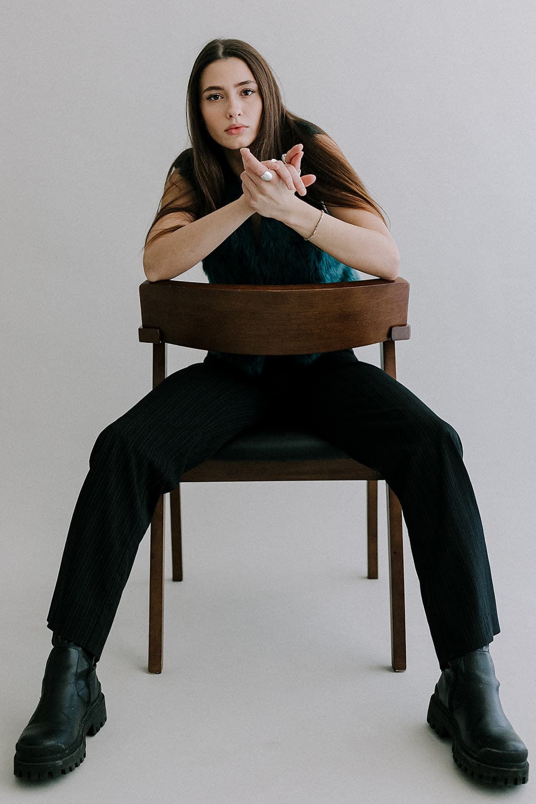 A model sits on a chair with hands clasped, staring directly into the camera, photographed in the natural light environment of Revelator Studio, Shreveport.