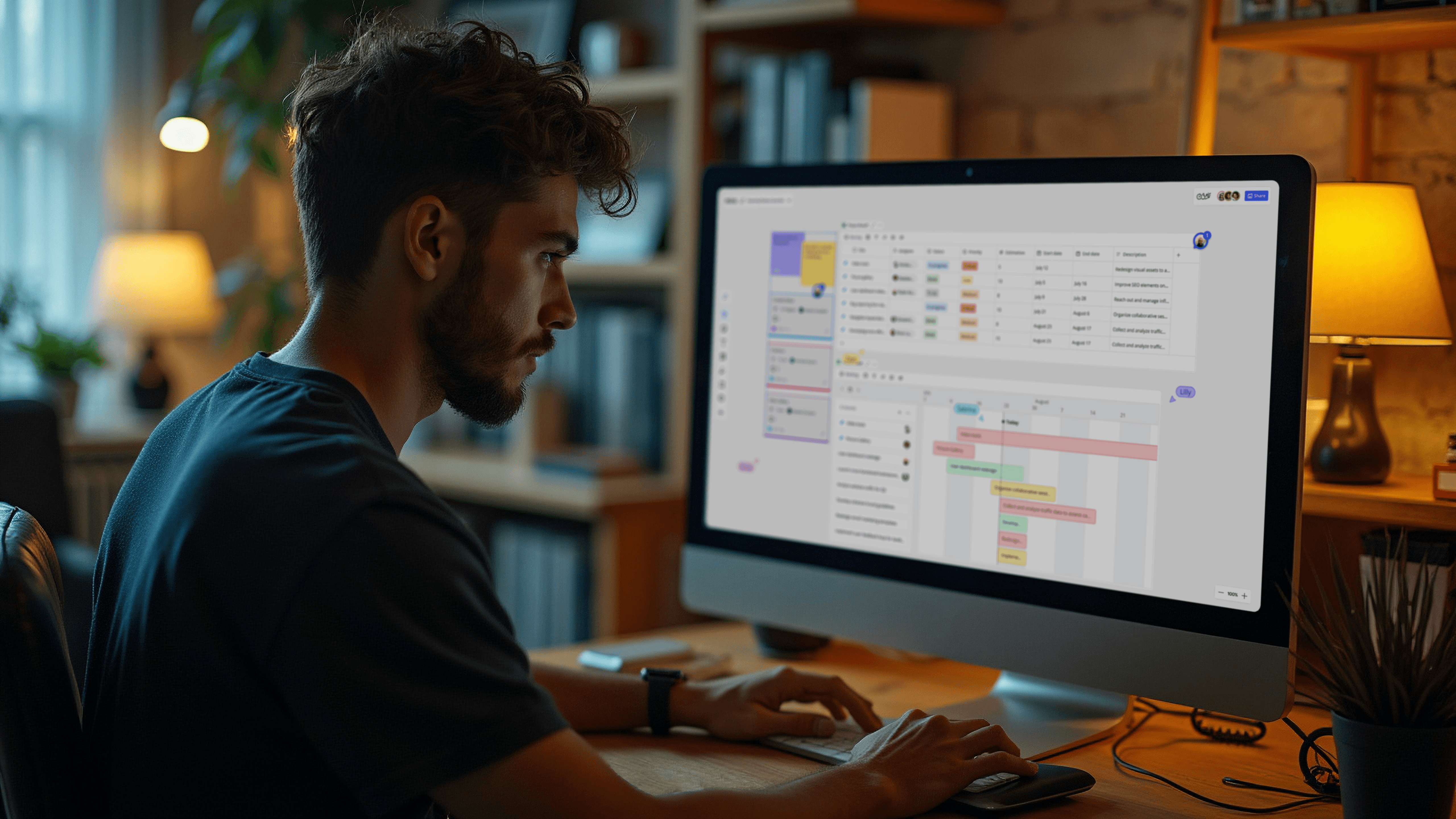 A man with dark hair and a beard sits at a wooden desk in a dimly lit room, focusing intently on a large computer screen displaying a colorful project management dashboard.