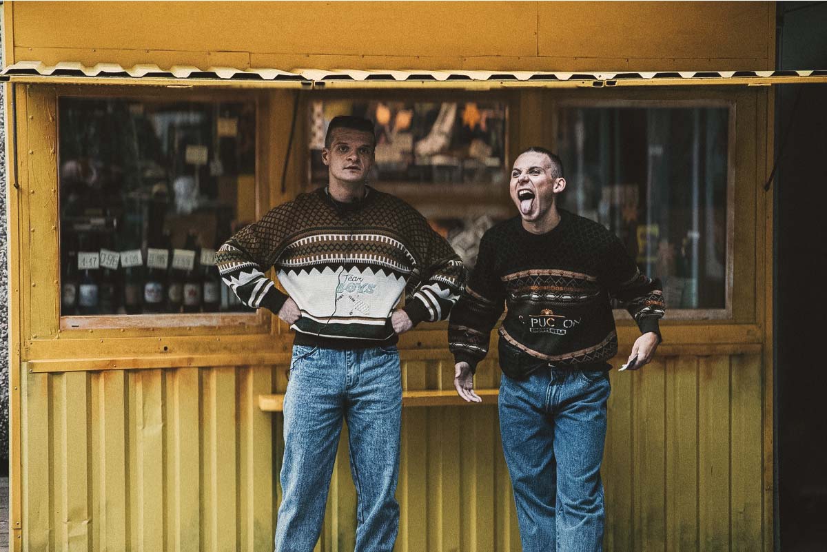 Rimants (Džiugas Grinys) and Mindė (Robertas Petraitis) standing near a street kiosk in Southern Chronicles, a 90s Baltic comedy-drama about friendship and rebellion.