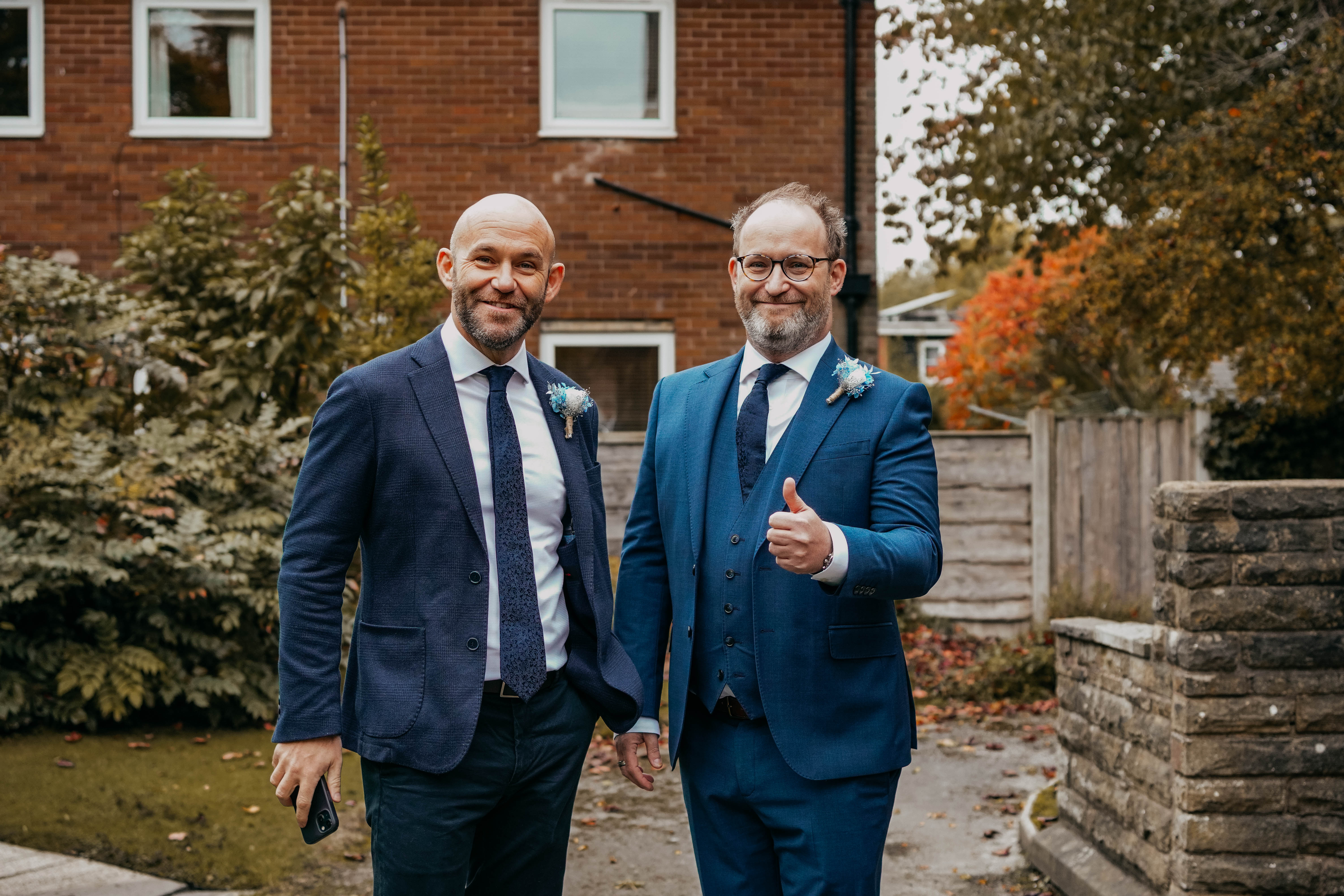 The groom, in a blue three-piece suit, gives a thumbs-up to the camera while standing next to his smiling best man. They are outdoors in front of a brick house with autumn foliage.