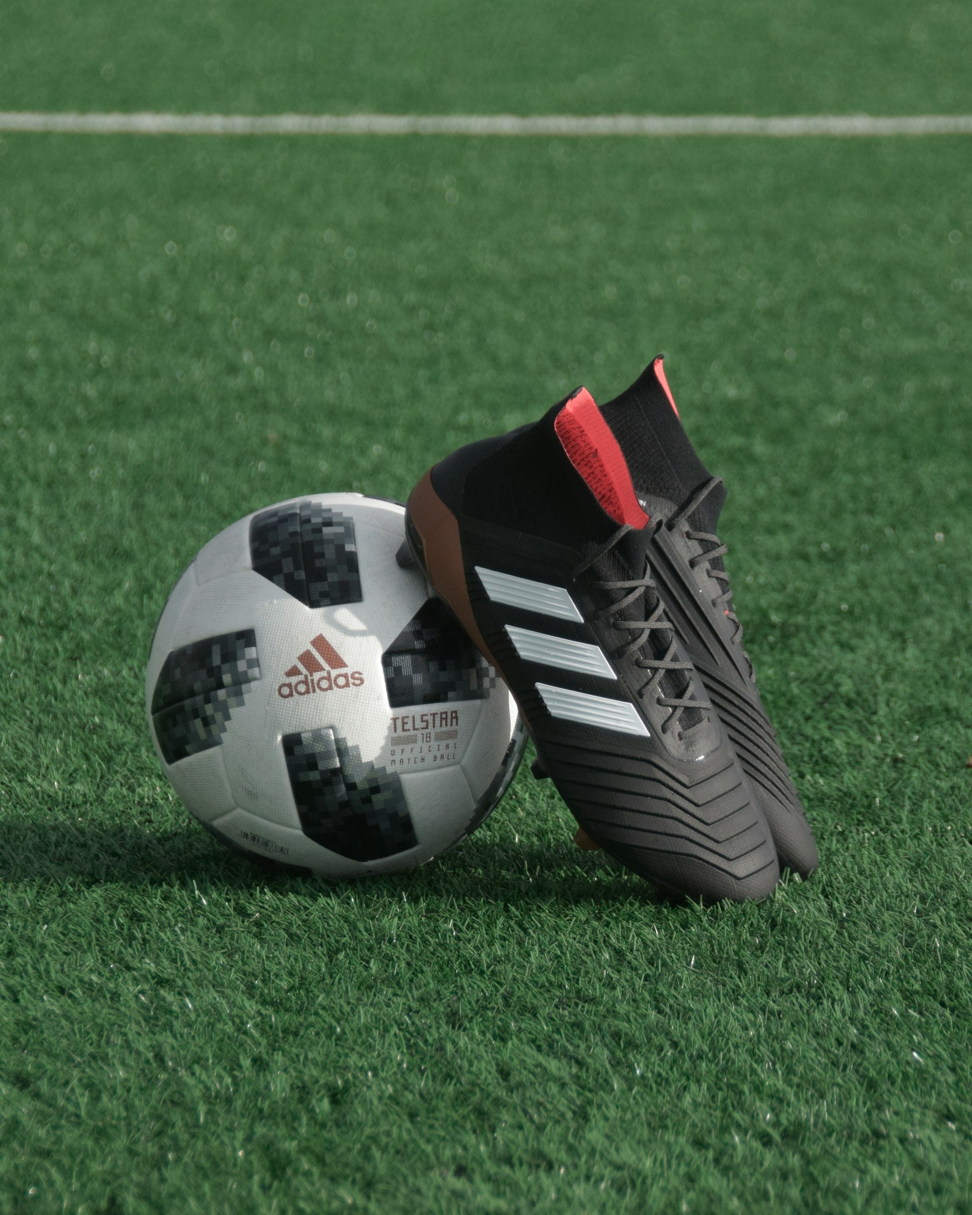 A pair of black Adidas soccer cleats resting against a white and black Telstar 18 soccer ball on green turf.