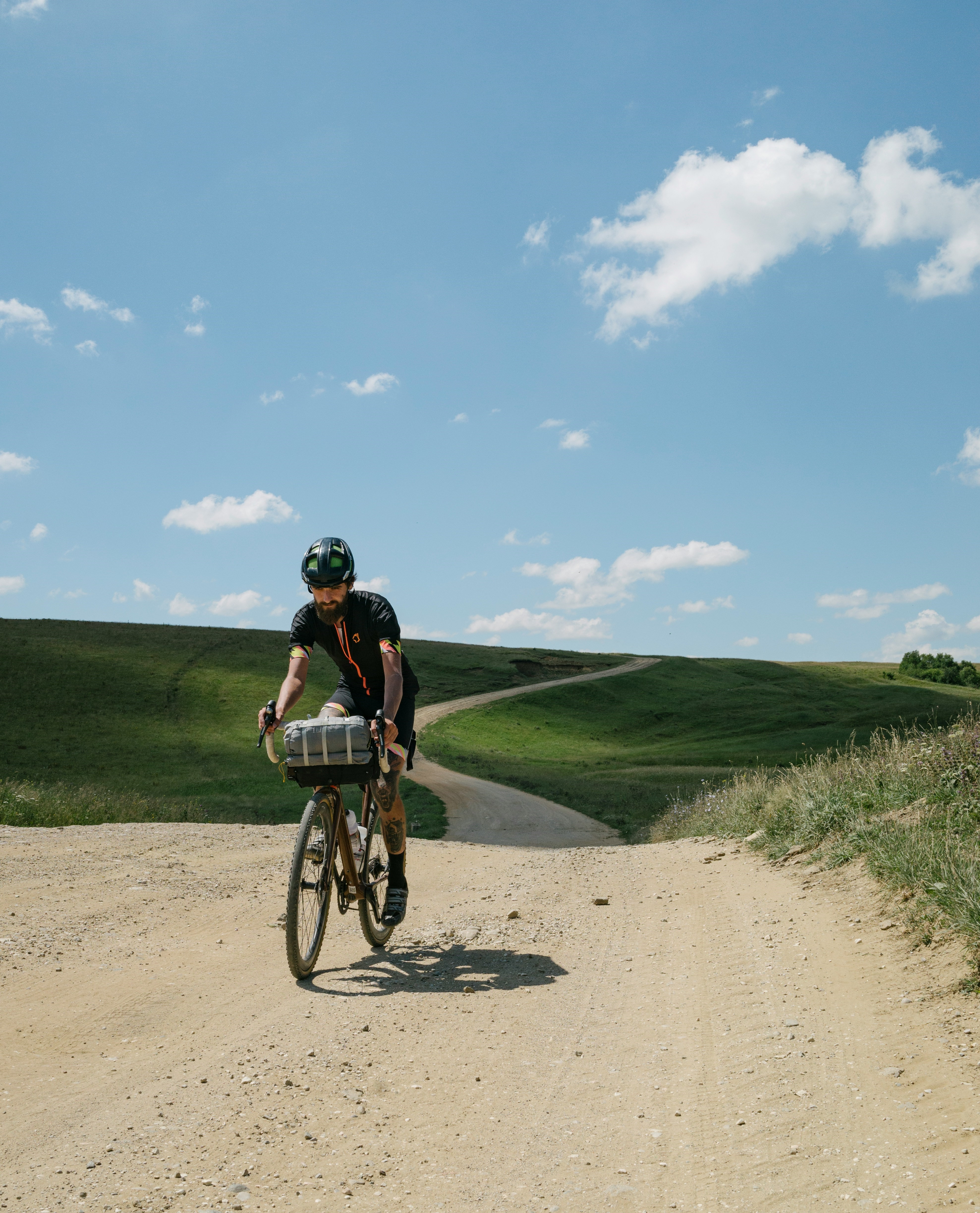 Cycliste pratiquant le vélo gravel sur un large chemin de terre poussiéreux en pleine nature sous le soleil.