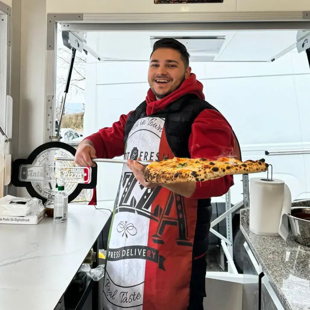 Freundlicher Pizzaiolo präsentiert frische Pizza auf Schaufel im Verkaufsanhänger Pizza Dielsdorf
