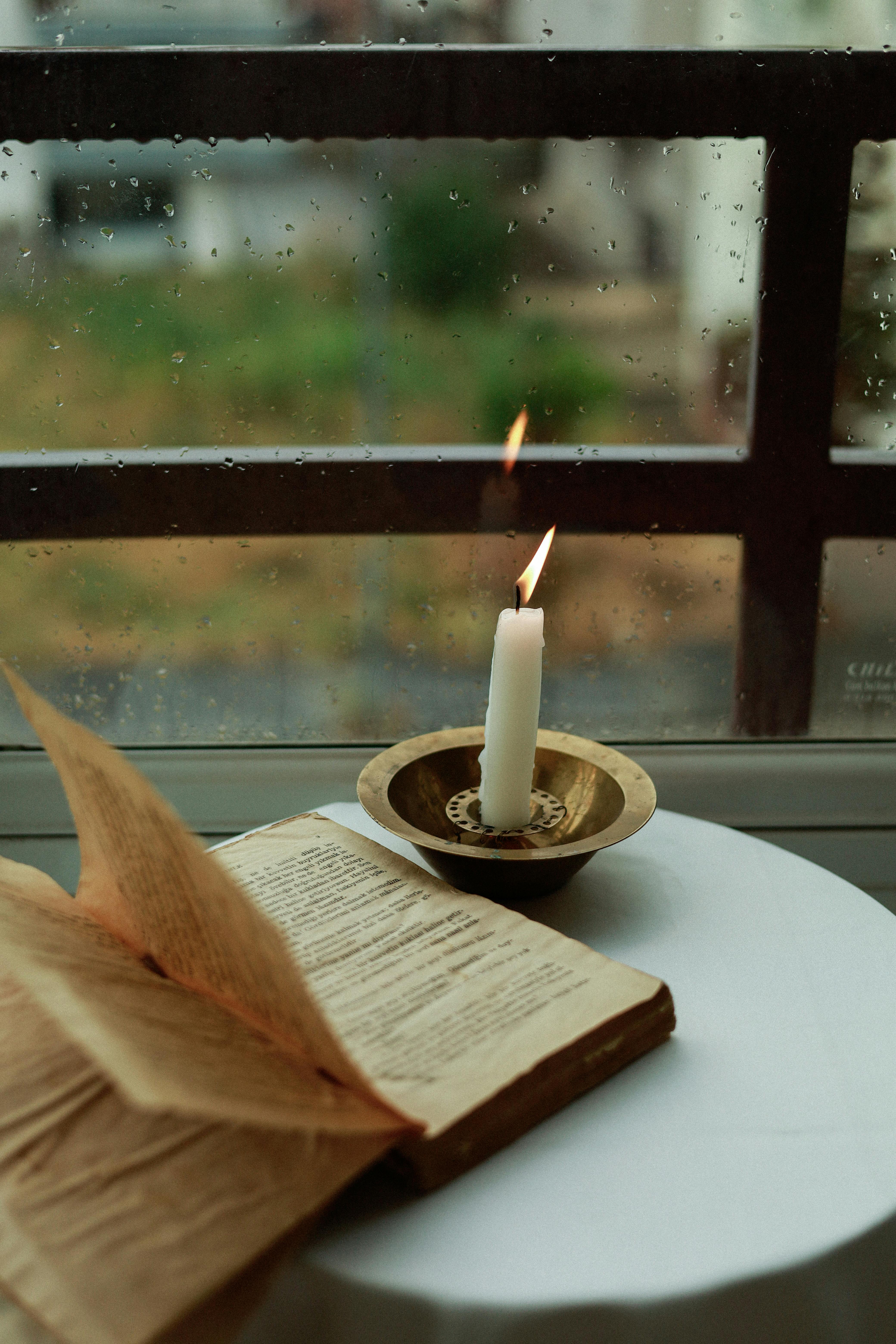 Lit candle beside an open book near a window in a calm setting