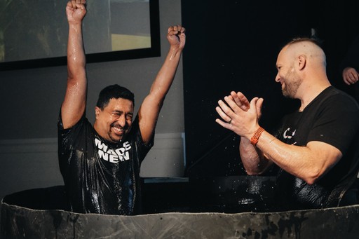 A group of people sitting on grass under trees, enjoying a sunny day near a body of water.