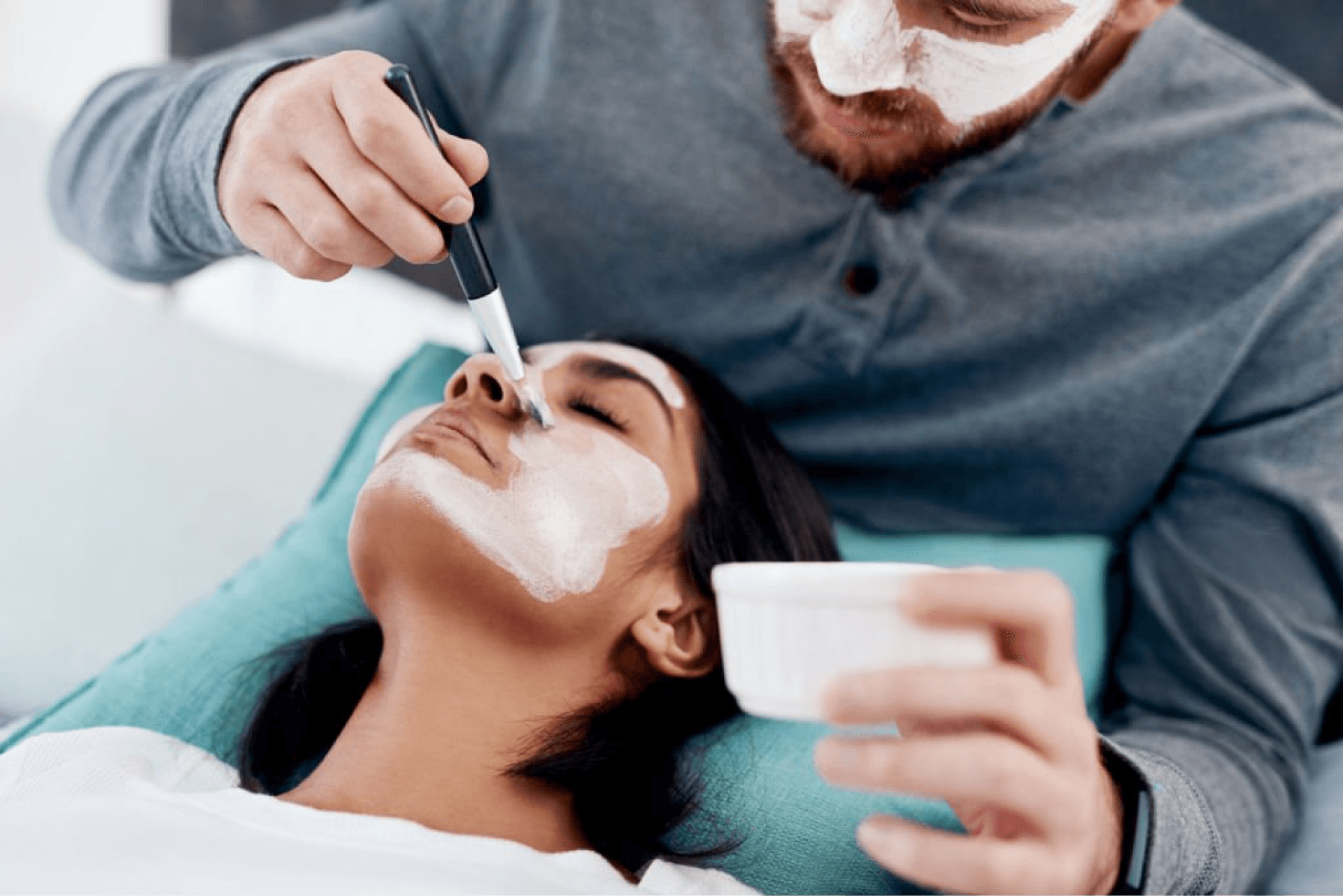 Man applying a face mask to his partner for wedding skin care rituals.