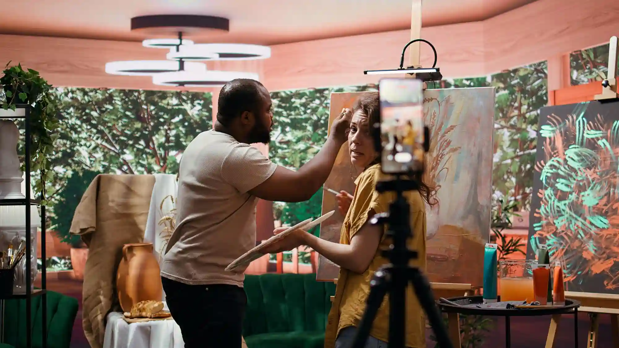 Two people interacting in an art studio, one possibly taking a video with a smartphone on a tripod.