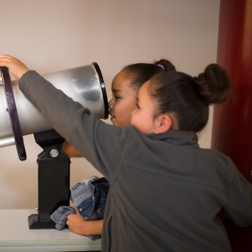 Dos niños miran a través de un gran telescopio plateado en interiores. Uno de los niños sostiene un pequeño juguete o una prenda de vestir.