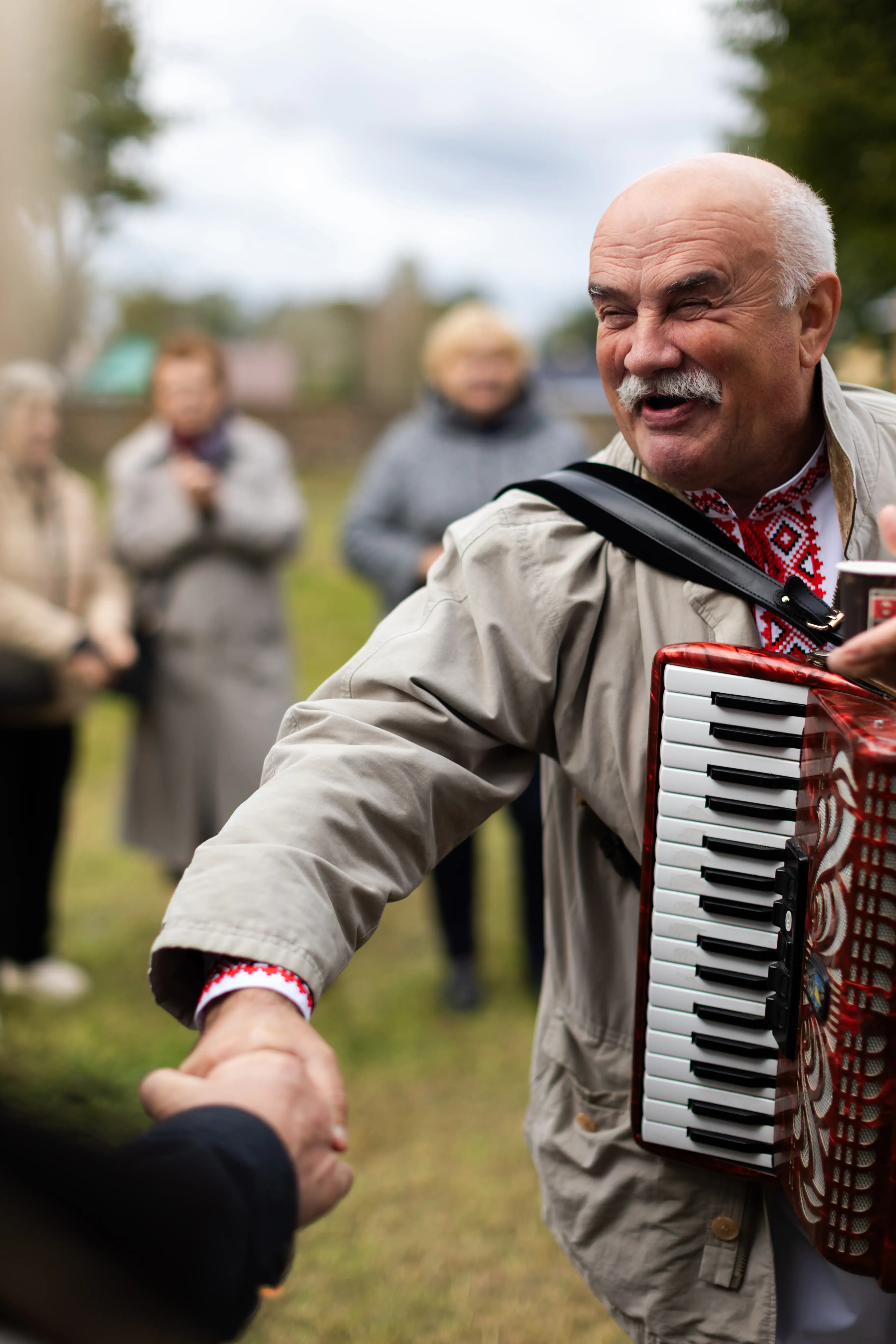 Starszy mężczyzna w jasnym płaszczu, grający na akordeonie, uśmiecha się i podaje rękę drugiej osobie. W tle widoczne postacie ludzi, którzy obserwują zdarzenie na świeżym powietrzu.