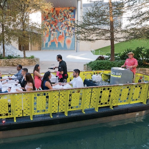 People dining on a yellow riverboat with a tour guide. Background includes trees, a colorful mural, and a modern building.