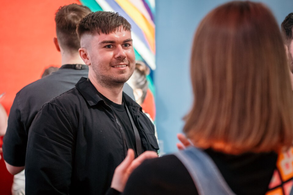 A group of people stand in conversation indoors. The foreground shows two individuals speaking to each other, with one facing the camera and the other facing away. The background includes others talking and a colourful wall with diagonal stripes.