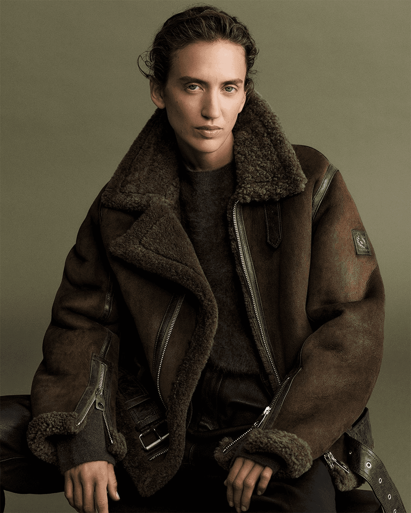 Dans le lookbook 2025 de la marque Belstaff, la mannequin porte un blouson type "aviateur" en peau lainée avec en fond un mur vert