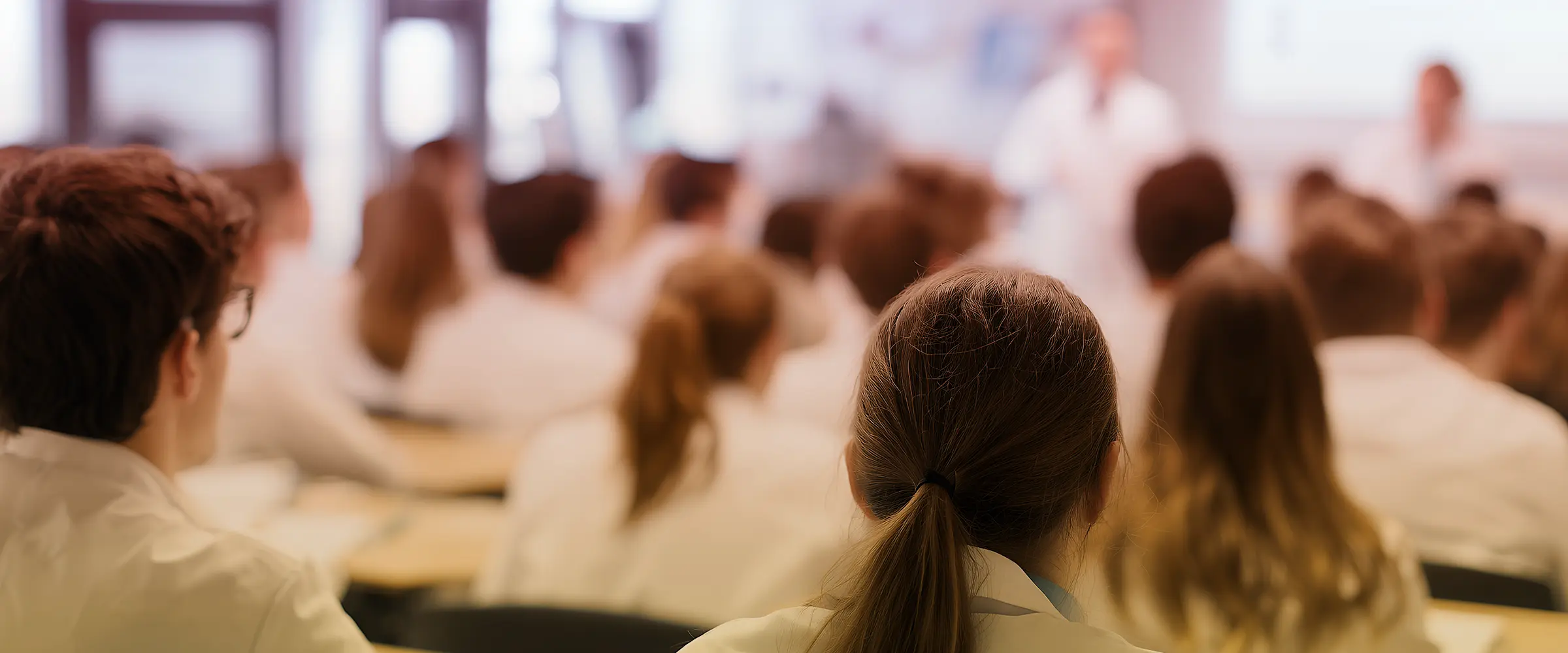 Students sitting in a classroom, viewed from behind, attentively listening to a teacher giving a lecture at the front of the room.