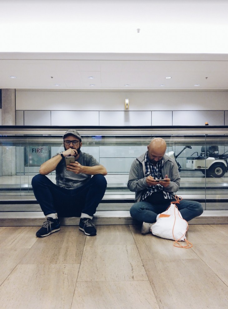 Dwóch mężczyzn siedzi na podłodze terminalu lotniska przed ruchomym chodnikiem; jeden pije, drugi patrzy w telefon.