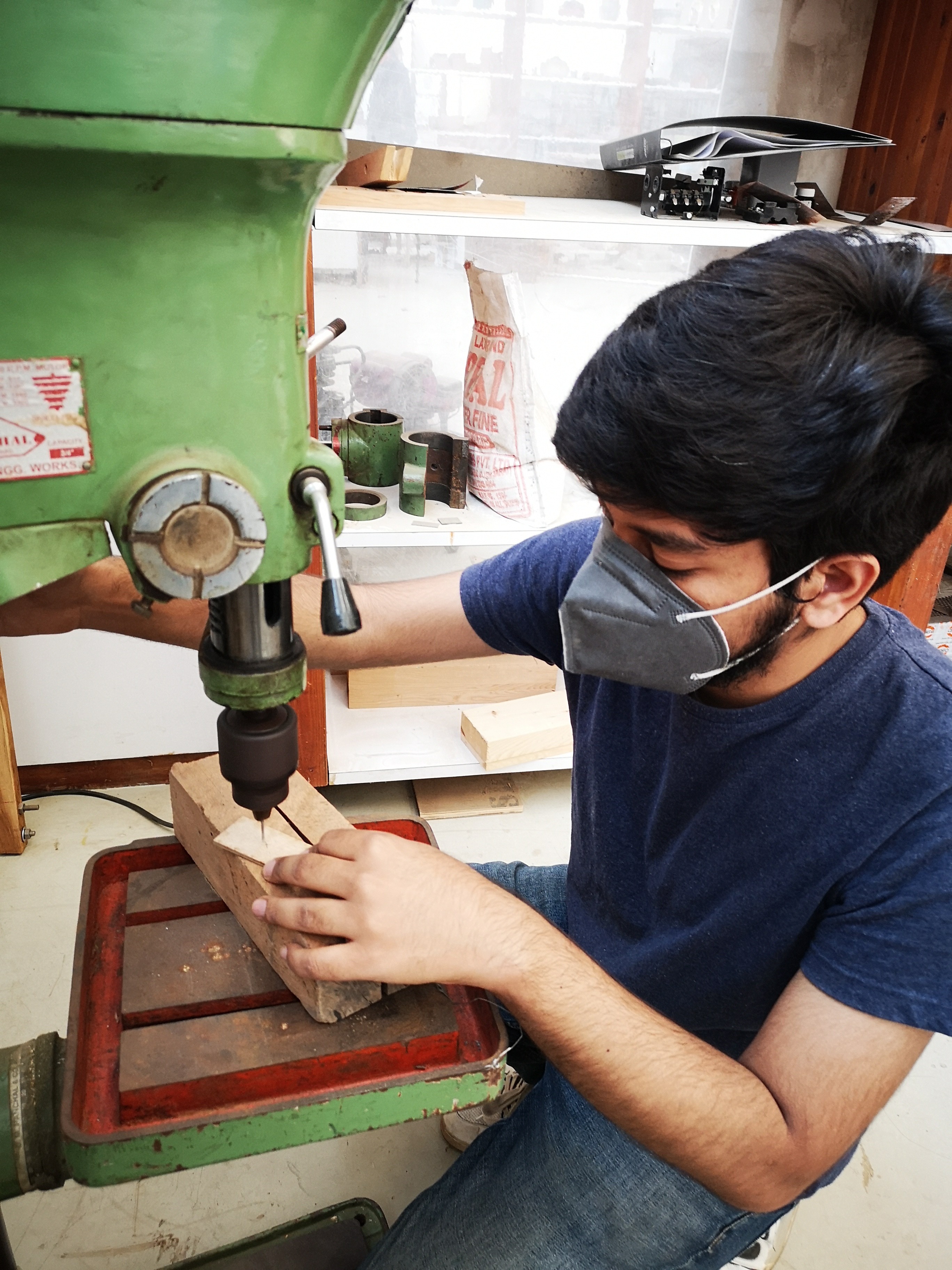 A spacious industrial-style interior with exposed brick walls, large windows, and natural light pouring in during sunset. Wooden desks with plants and workspace materials are arranged along the walls, with a view of greenery and an outdoor seating area.