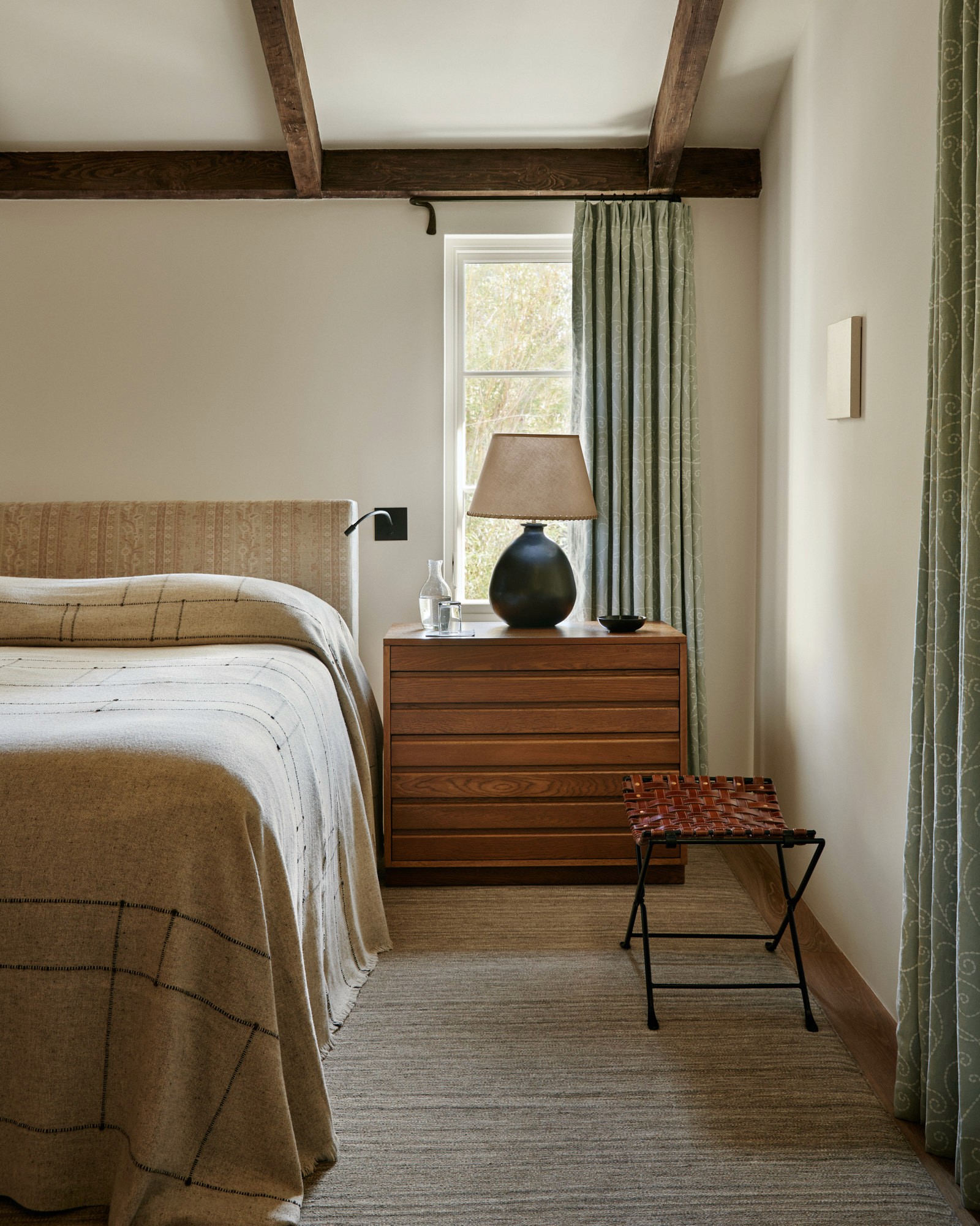 A bedroom with exposed ceiling beams, a bed dressed in a handwoven windowpane throw, and a slatted wood dresser topped with a dark ceramic lamp. A woven leather luggage rack sits nearby, and green patterned curtains frame the window.