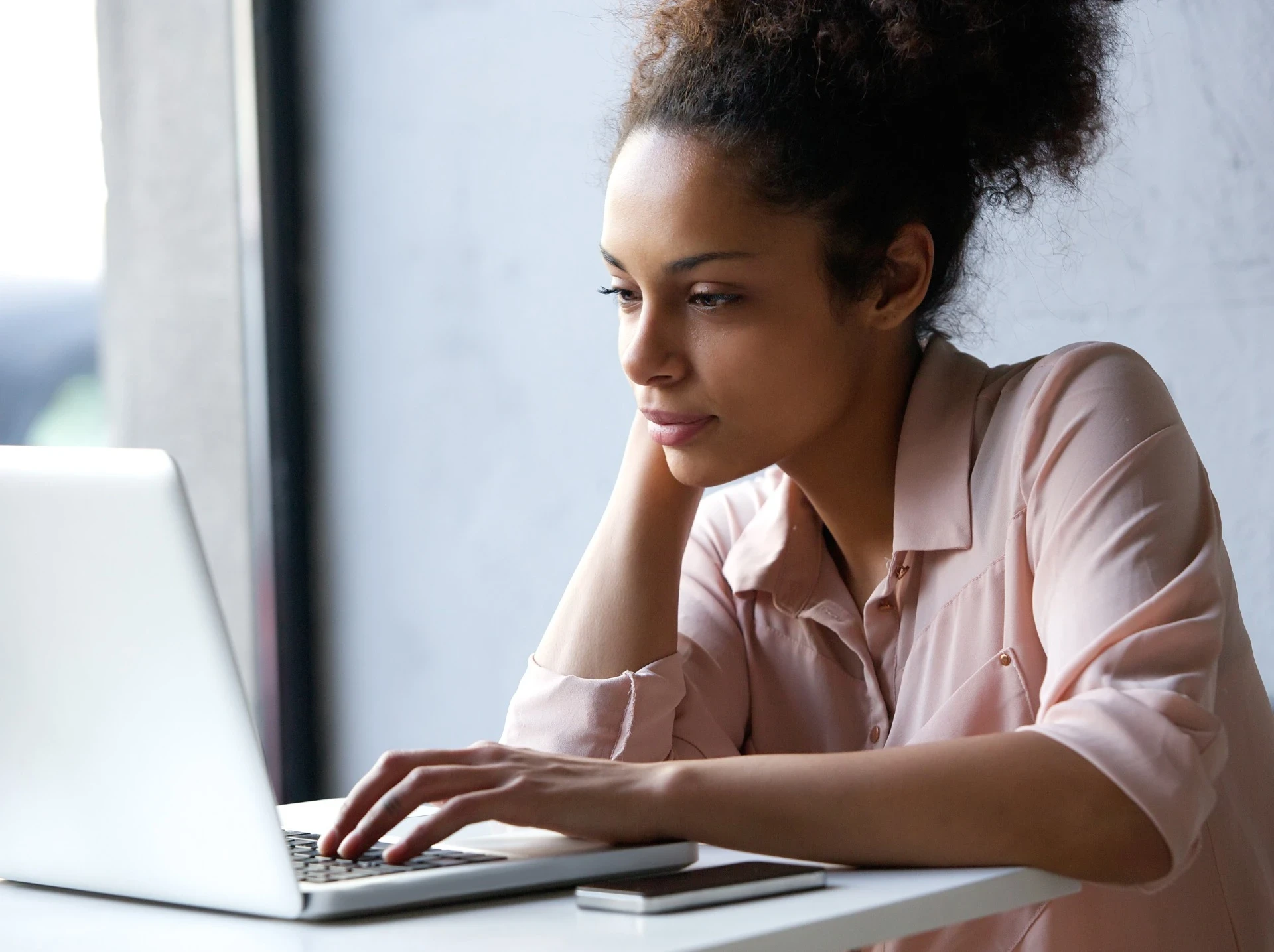 Imagem de uma mulher jovem com expressão calma e concentrada, olhando para a tela de um laptop. Ela apoia a cabeça com uma das mãos e usa a outra para digitar. Veste uma blusa clara, e o ambiente parece iluminado pela luz natural que entra pela janela ao lado. No canto da mesa, um celular está posicionado ao lado do laptop. A imagem transmite um momento de foco e produtividade, representando o trabalho ou estudo em um ambiente tranquilo e confortável.