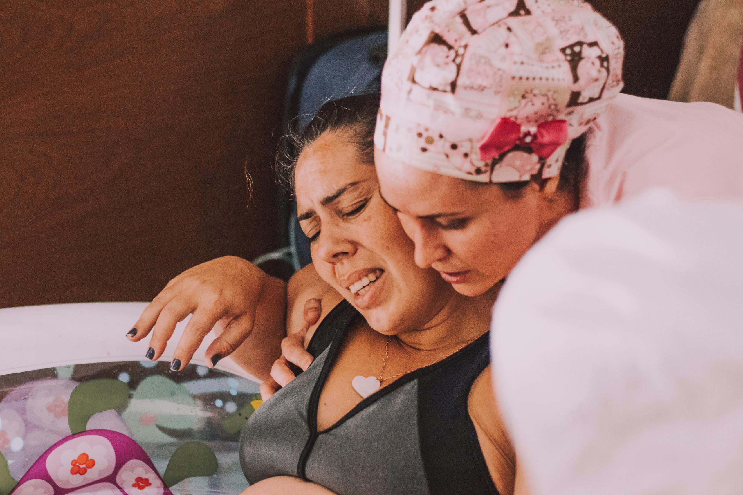 A woman is sitting in a birthing pool, she is concentrating and having a contraction. She is being held up and supported by a medical practioner in a pattern scrub hat.