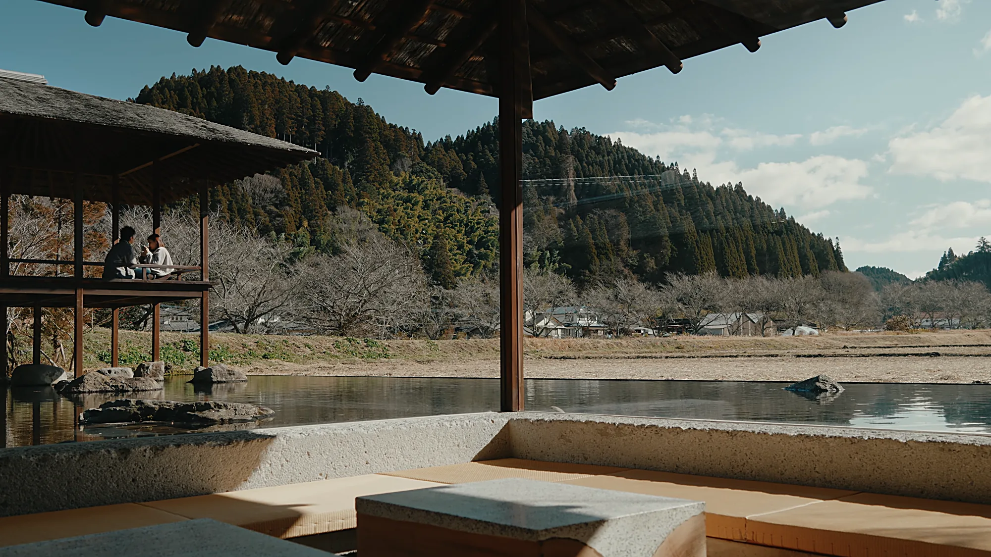 A thatched open pavilion over a pond near Kurokawa Onsen, two people seated, hills beyond