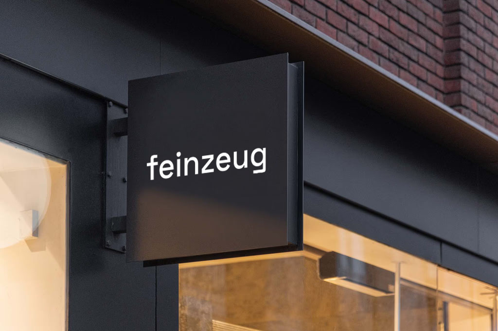 Modern architectural workspace with large floor-to-ceiling windows, surrounded by trees, and desks filled with design materials and books. Two blurred figures are walking through the space.