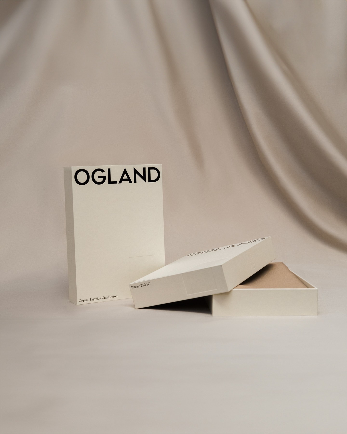 Cream Ogland bedding boxes with a large black wordmark and small product details, arranged open and closed against softly draped fabric.