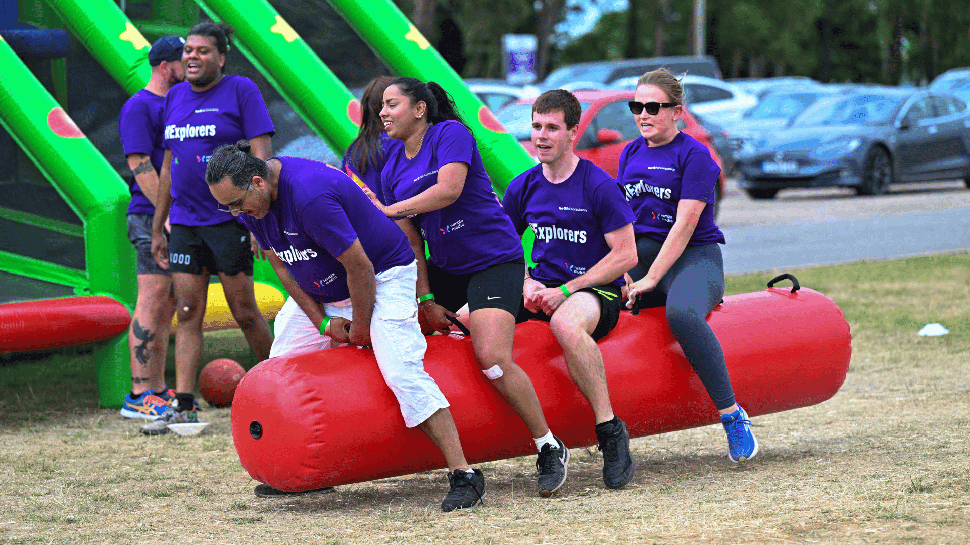 Outdoor team-building event with participants wearing matching blue shirts, taking part in inflatable obstacle courses and games, smiling and cheering, with the words 'ENTER A TEAM' in large bold white and green text across the image.