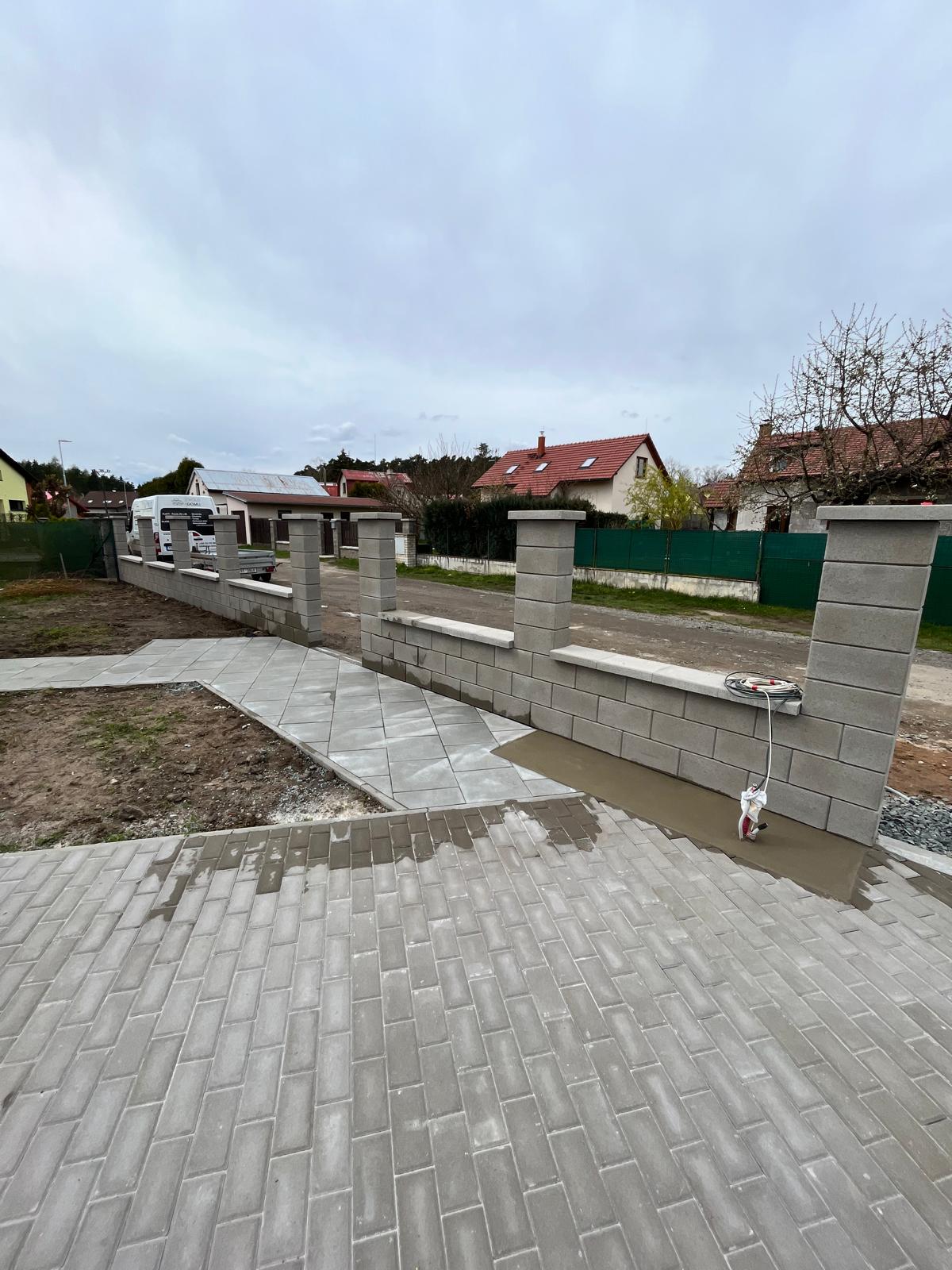Nová šedá zděná zeď s pilíři a chodník z dlažebních kostek. Stavební práce, dům v pozadí.