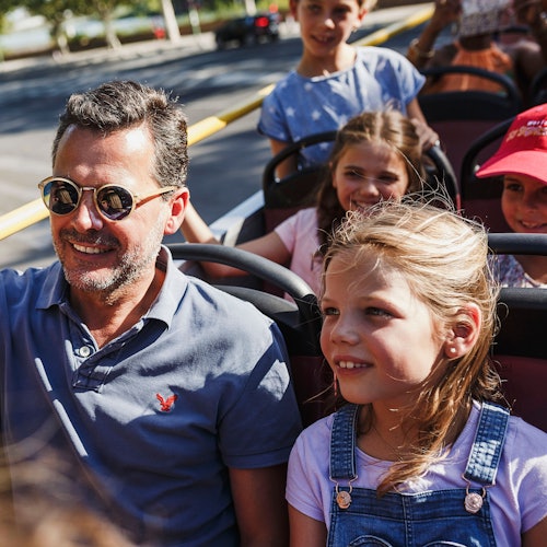 A man and four children smiling and sitting on a sunny open-top bus ride.