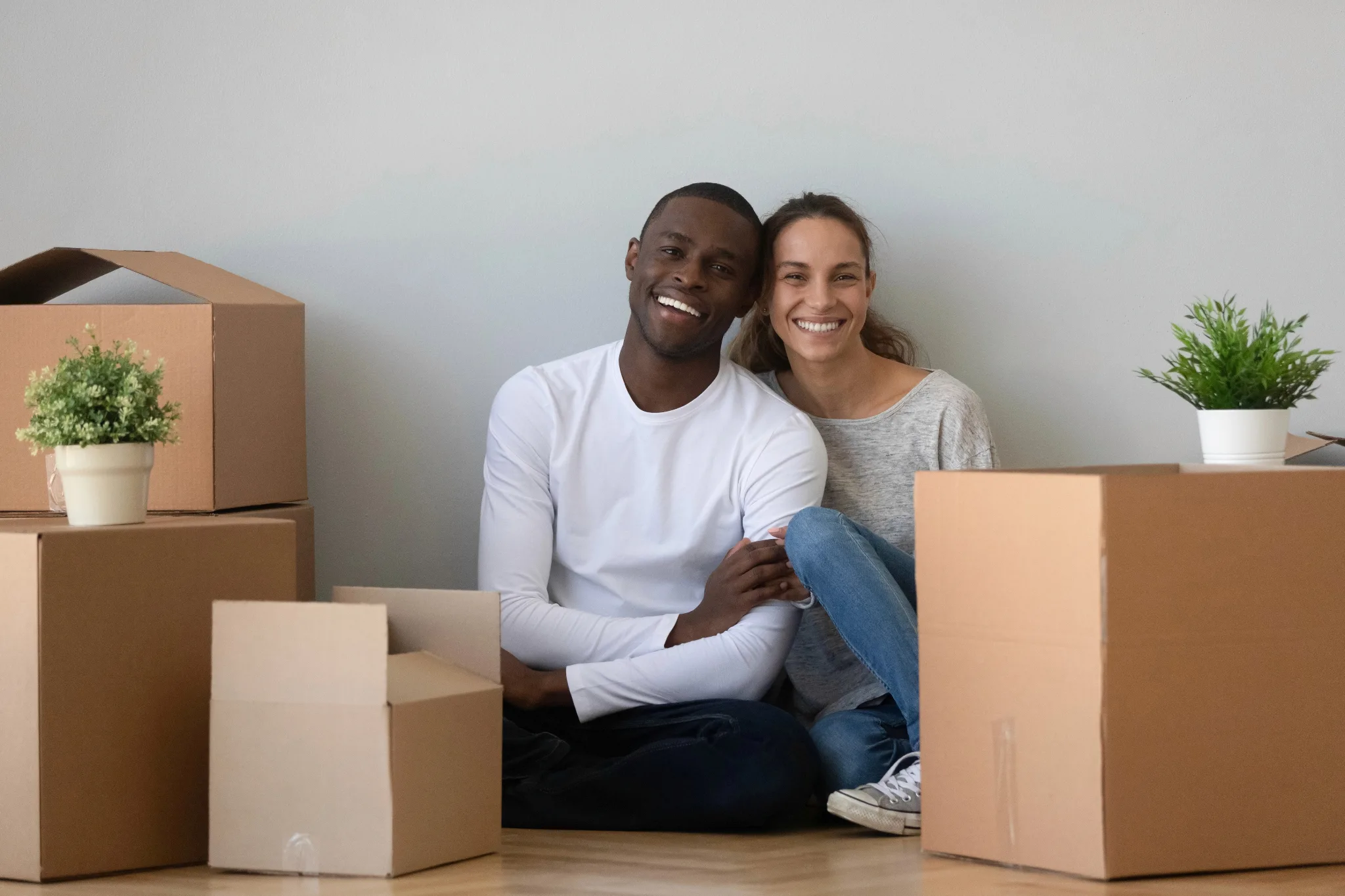 A smiling young couple, sitting on the floor surrounded by moving boxes.