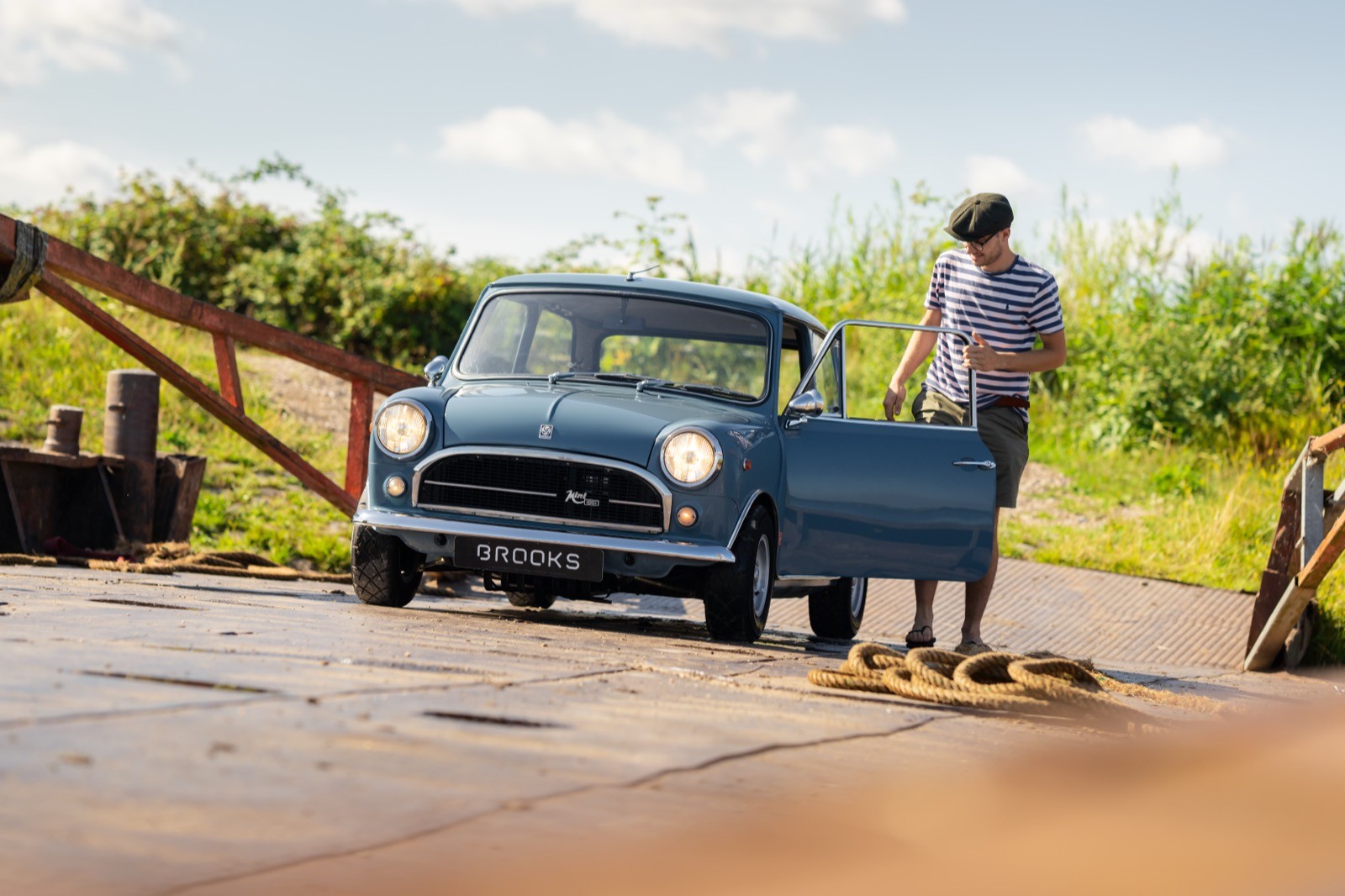 A person stands next to a blue car on a muddy road, with green scenery in the background.